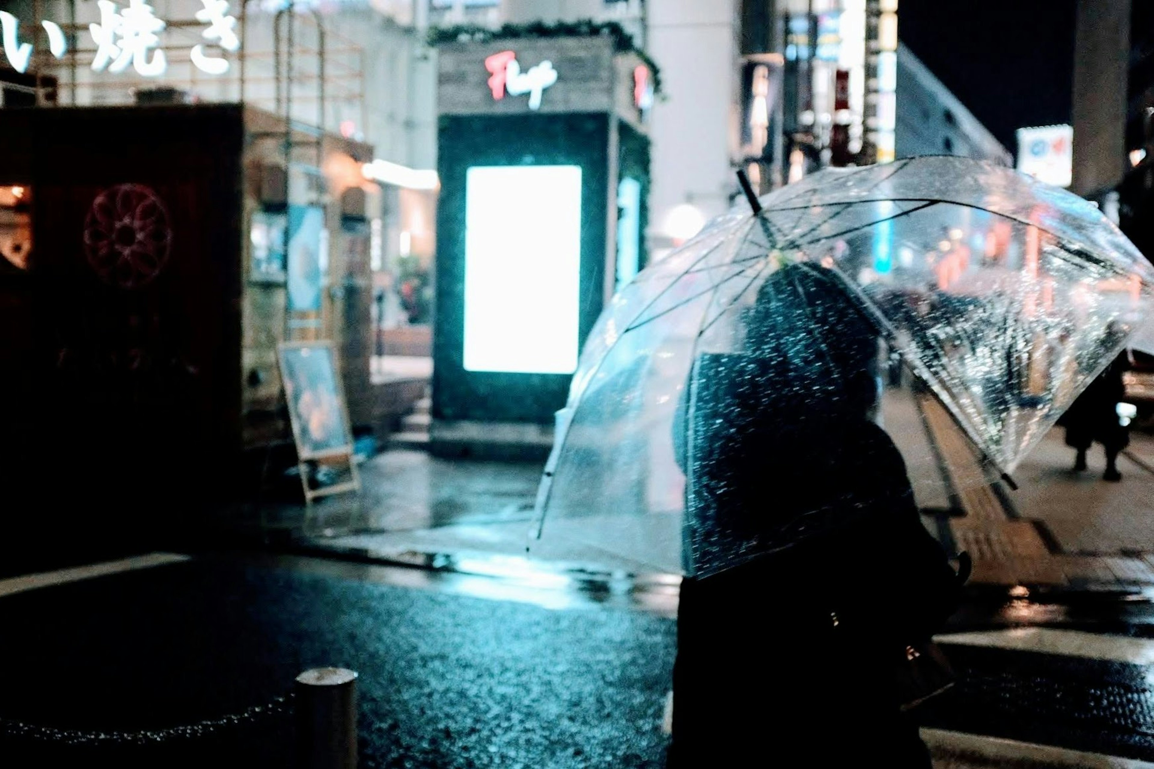 A person holding a transparent umbrella in the rain with city lights in the background at night