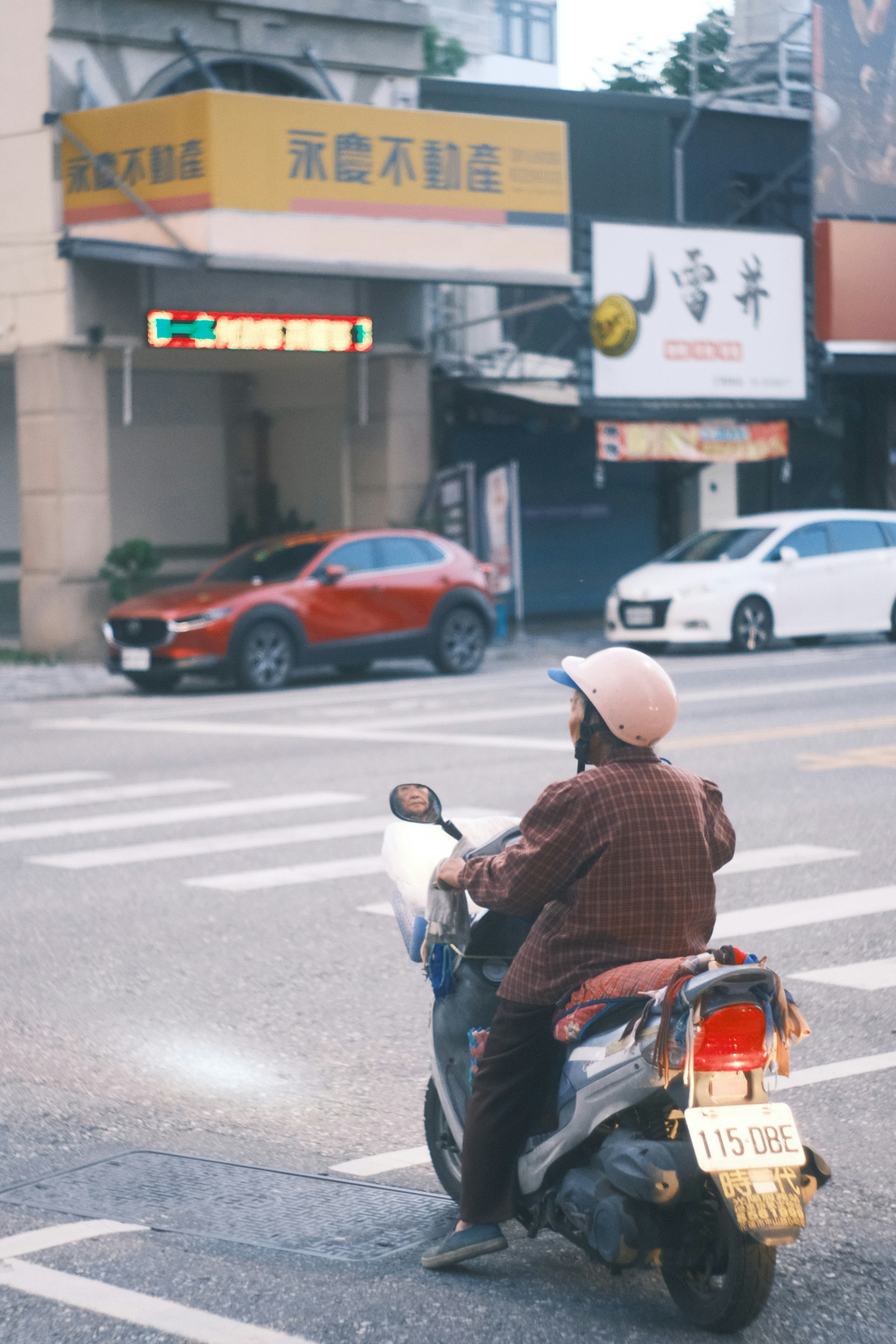 A person on a scooter crossing the street with storefronts and cars in view