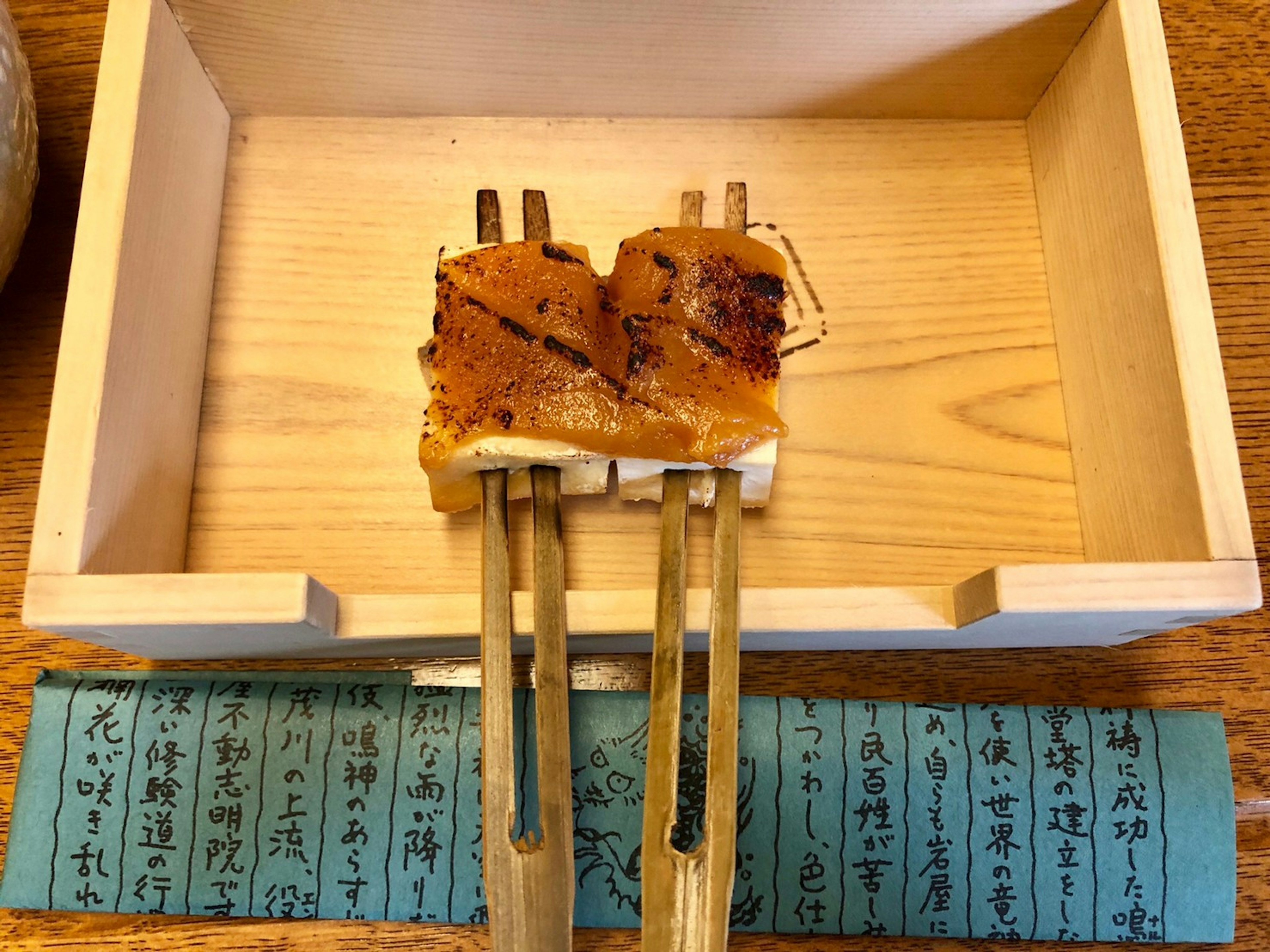 Grilled fish served on chopsticks above a wooden box with blue paper