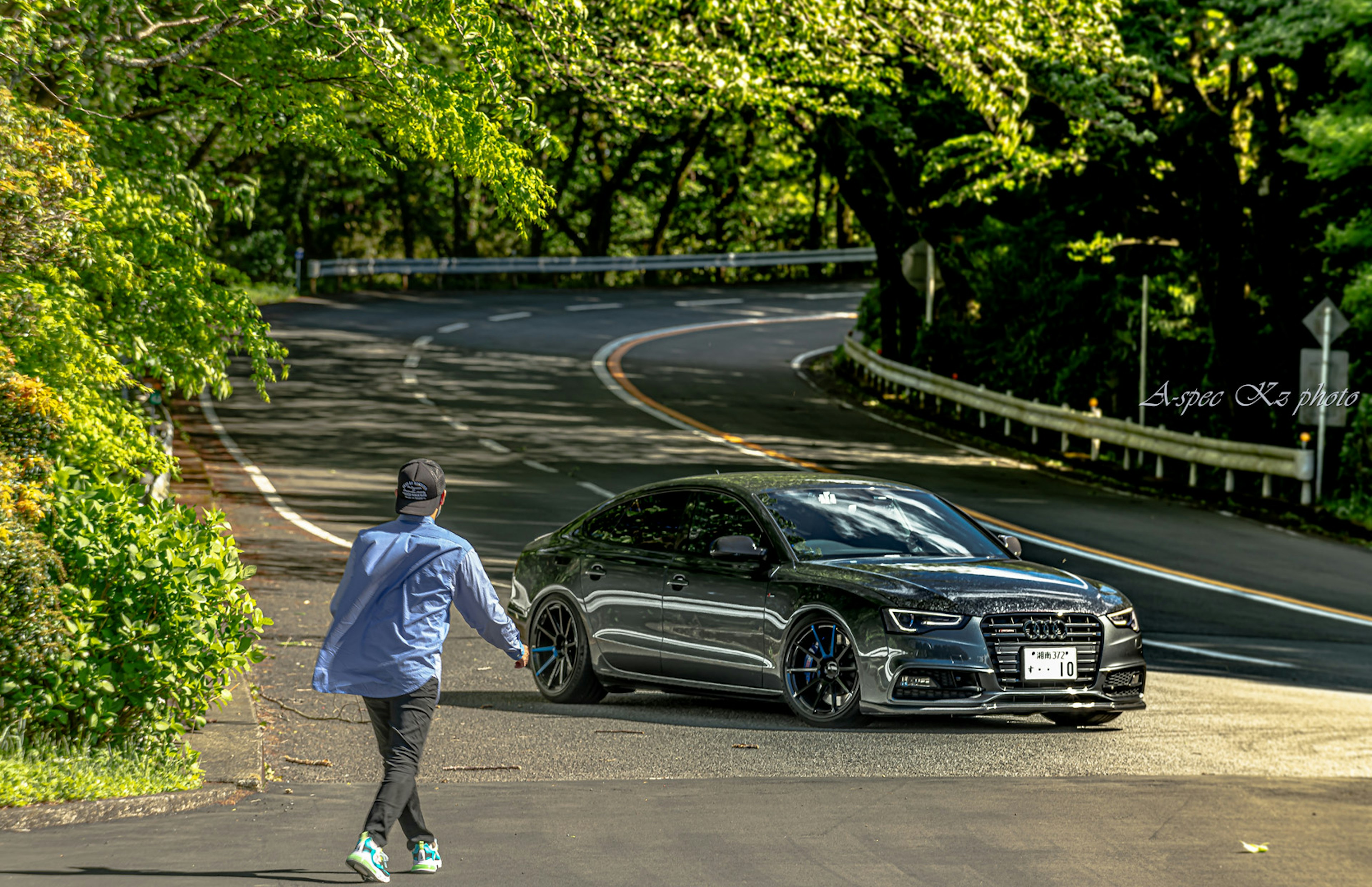 一個人在郁郁蔥蔥的道路上走，旁邊是一輛黑色跑車