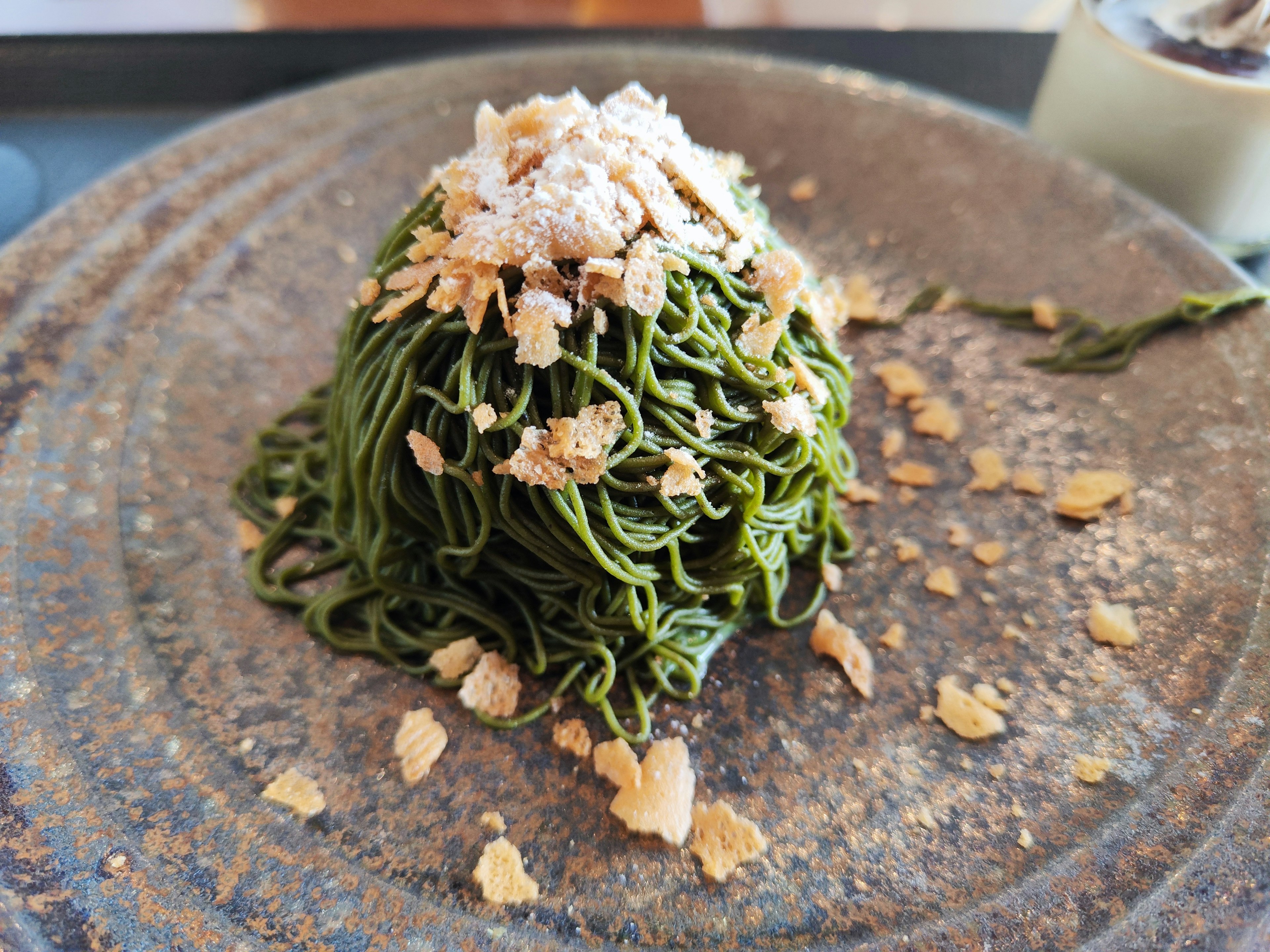 A mound of green noodles topped with crunchy flakes on a textured plate