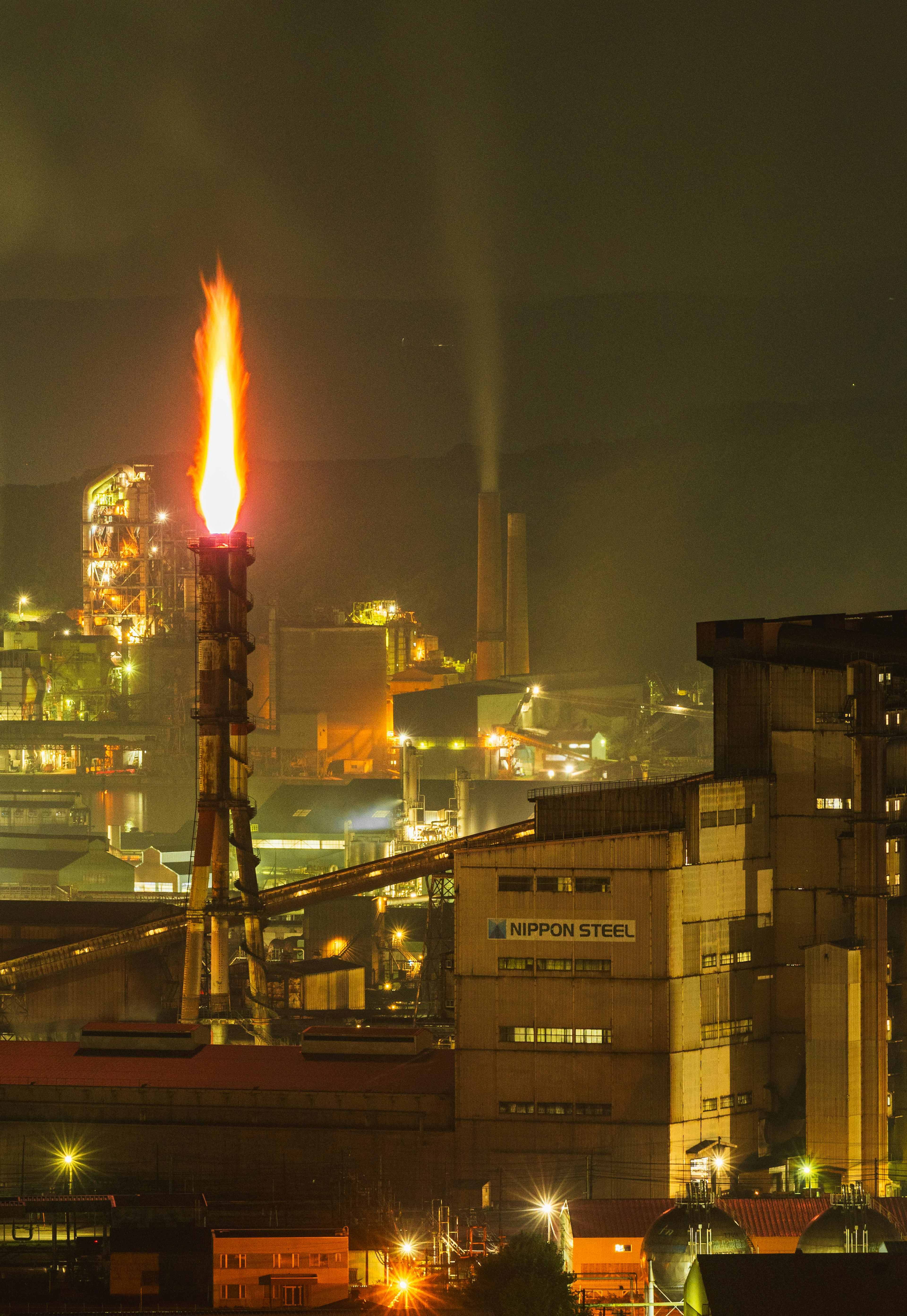 Industrial landscape at night featuring a flaming smokestack and factories