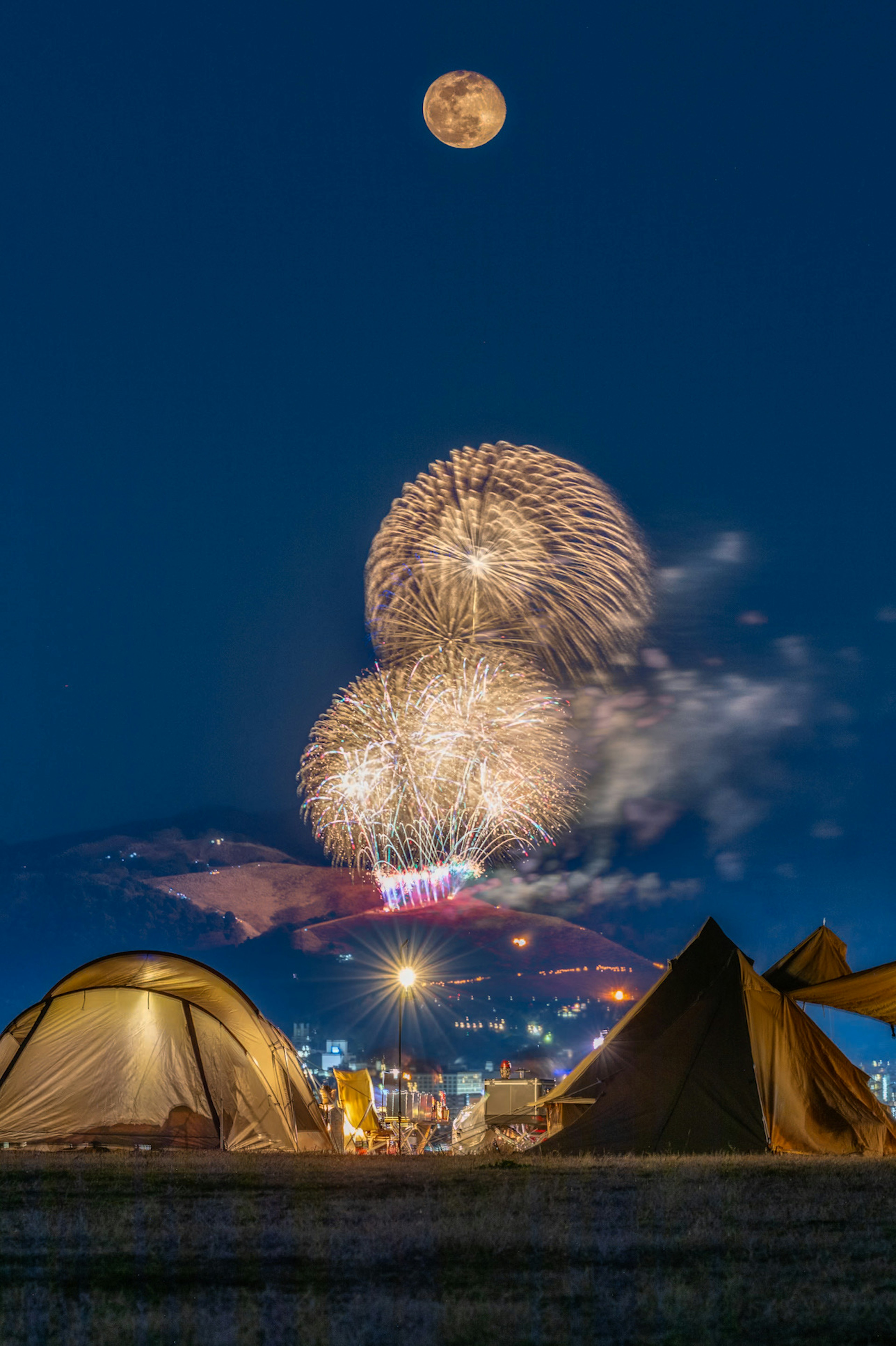 夜空に輝く花火と満月の下にテントが並ぶ風景