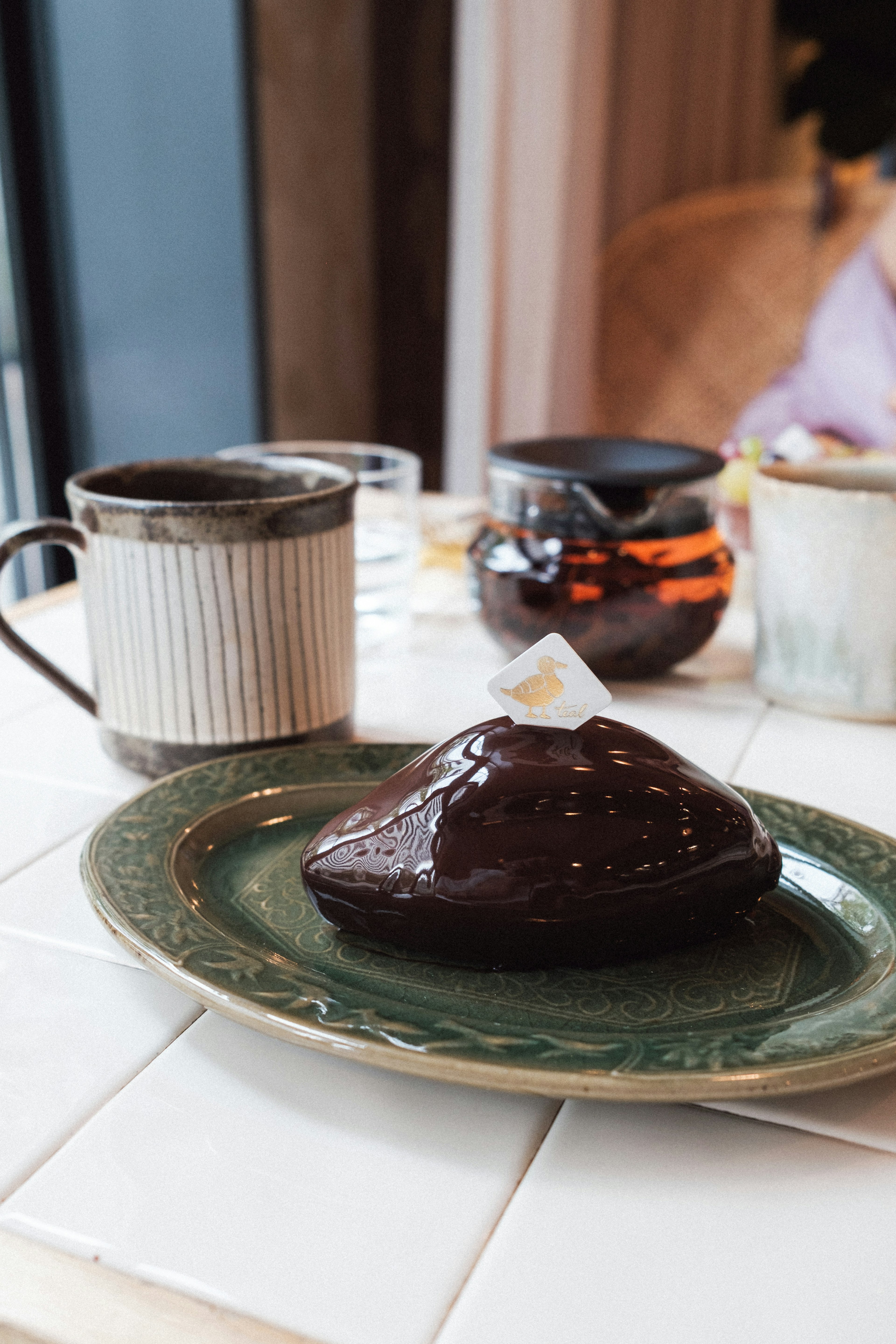 Pastel de chocolate en un plato verde con taza de café y tetera al fondo