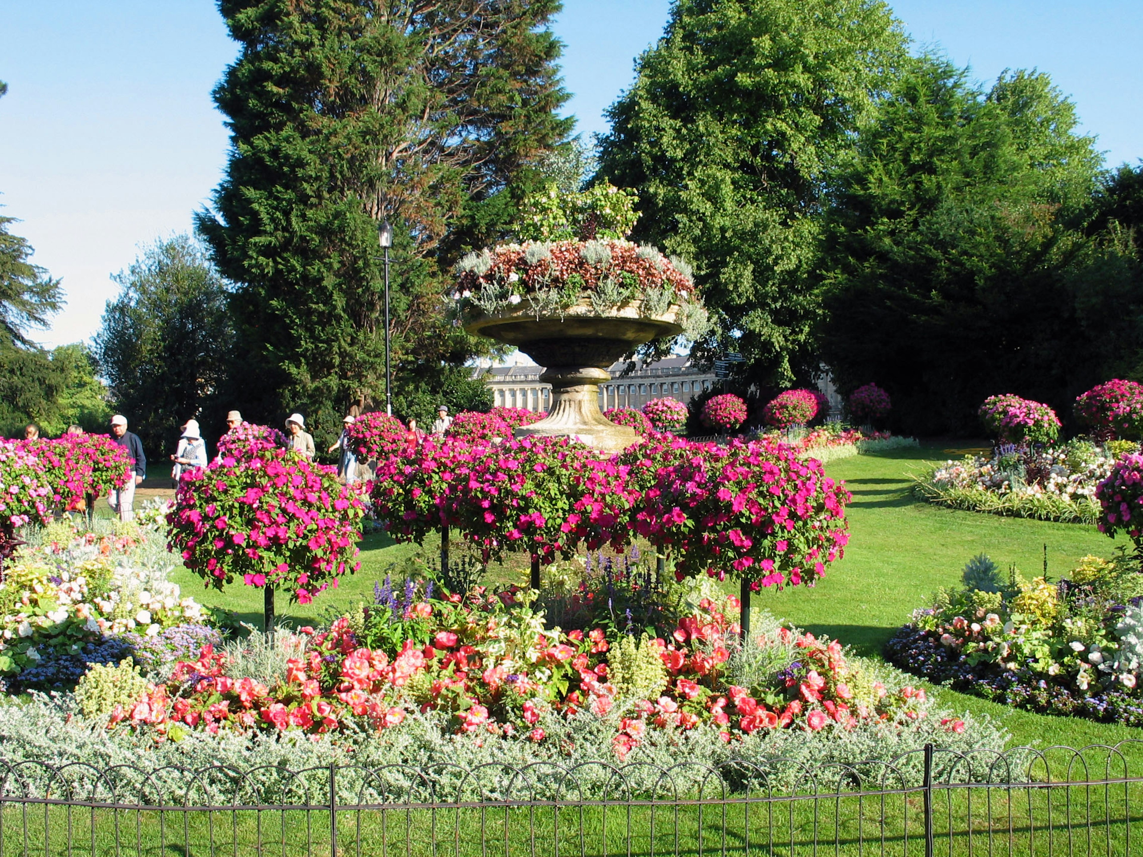 A beautiful garden featuring a large fountain surrounded by colorful flowers and greenery with visitors in the background