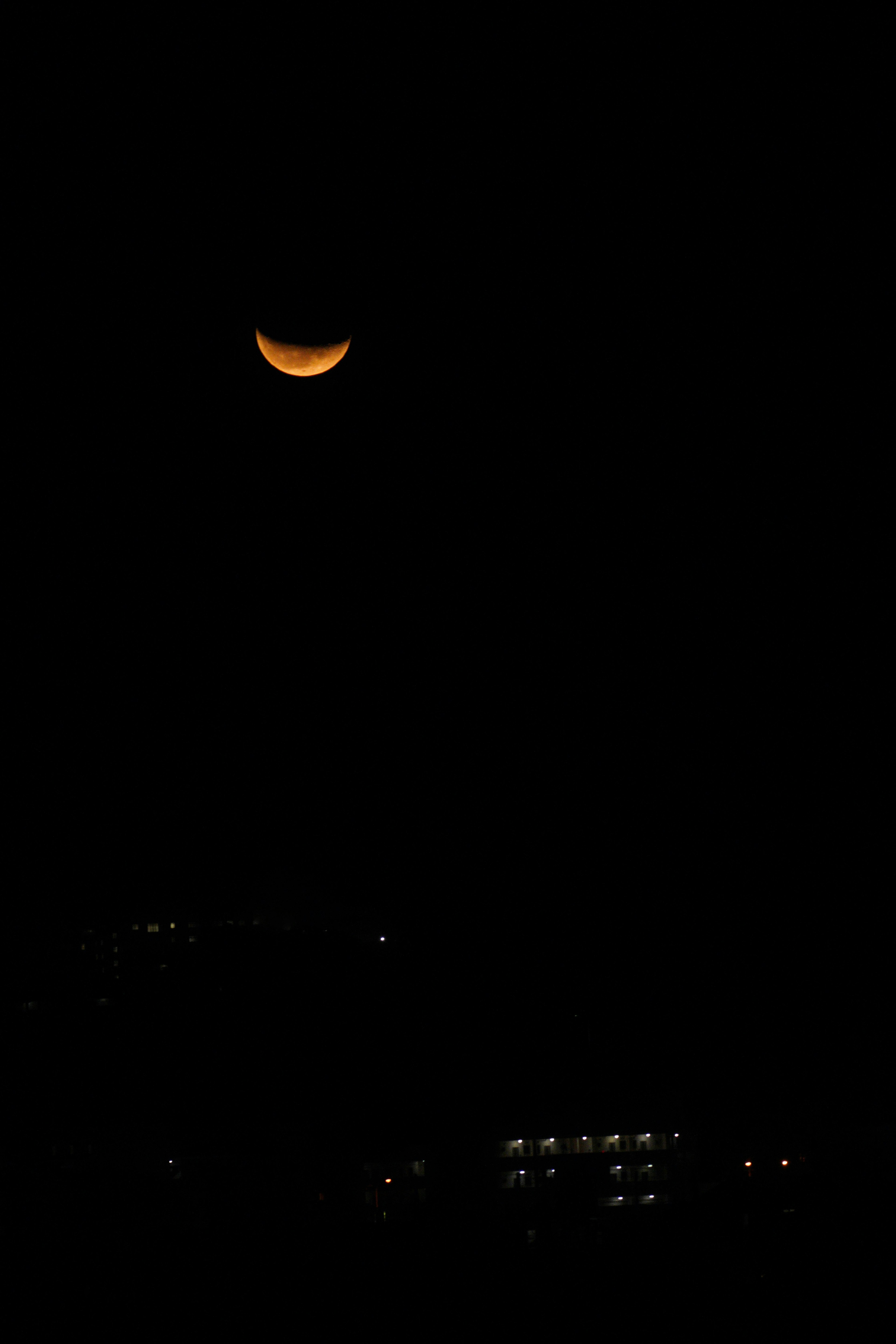 Luna creciente naranja en el cielo nocturno con luces tenues abajo