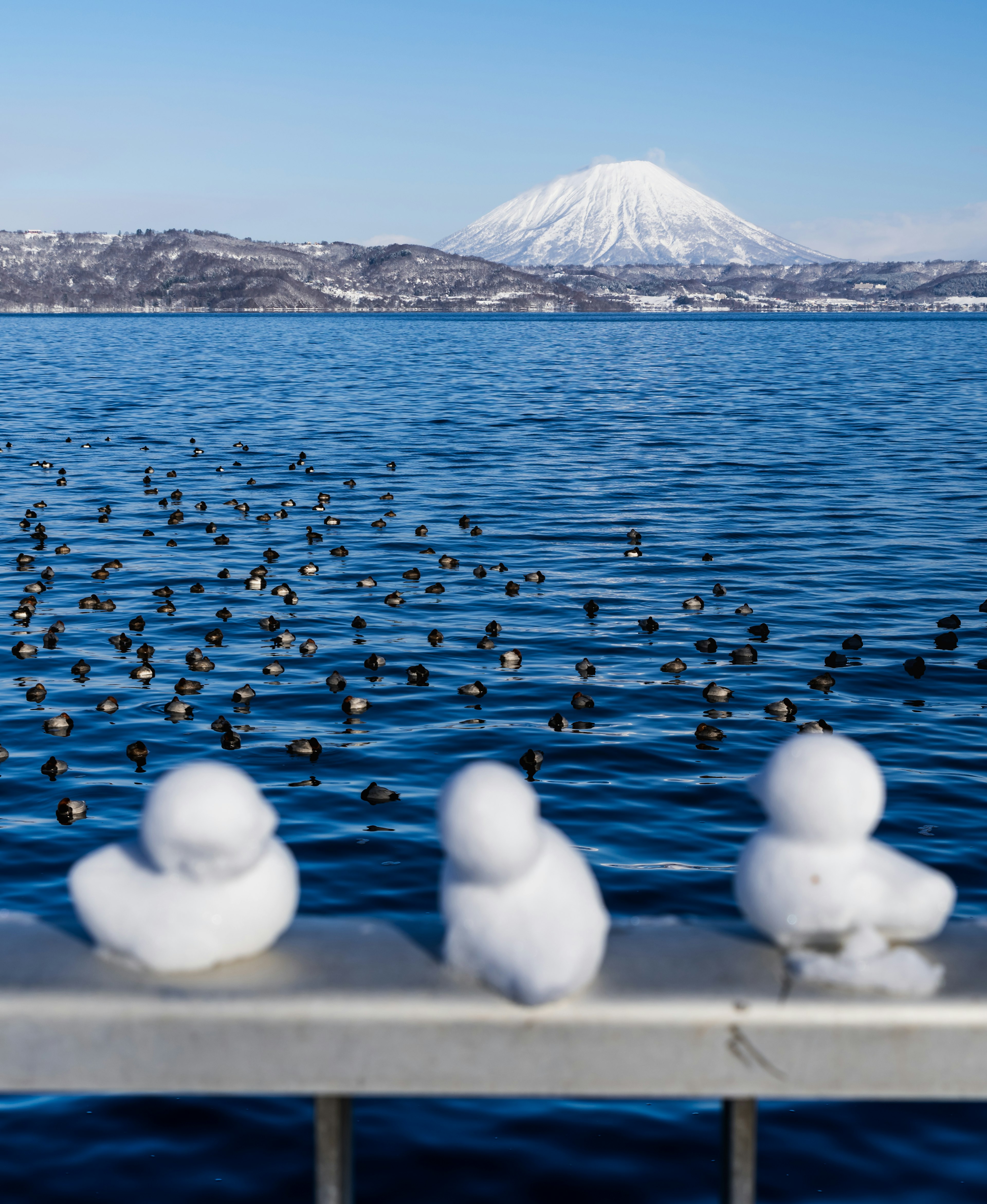 Three small snow figures lined up by a lake with a snow-capped mountain in the background