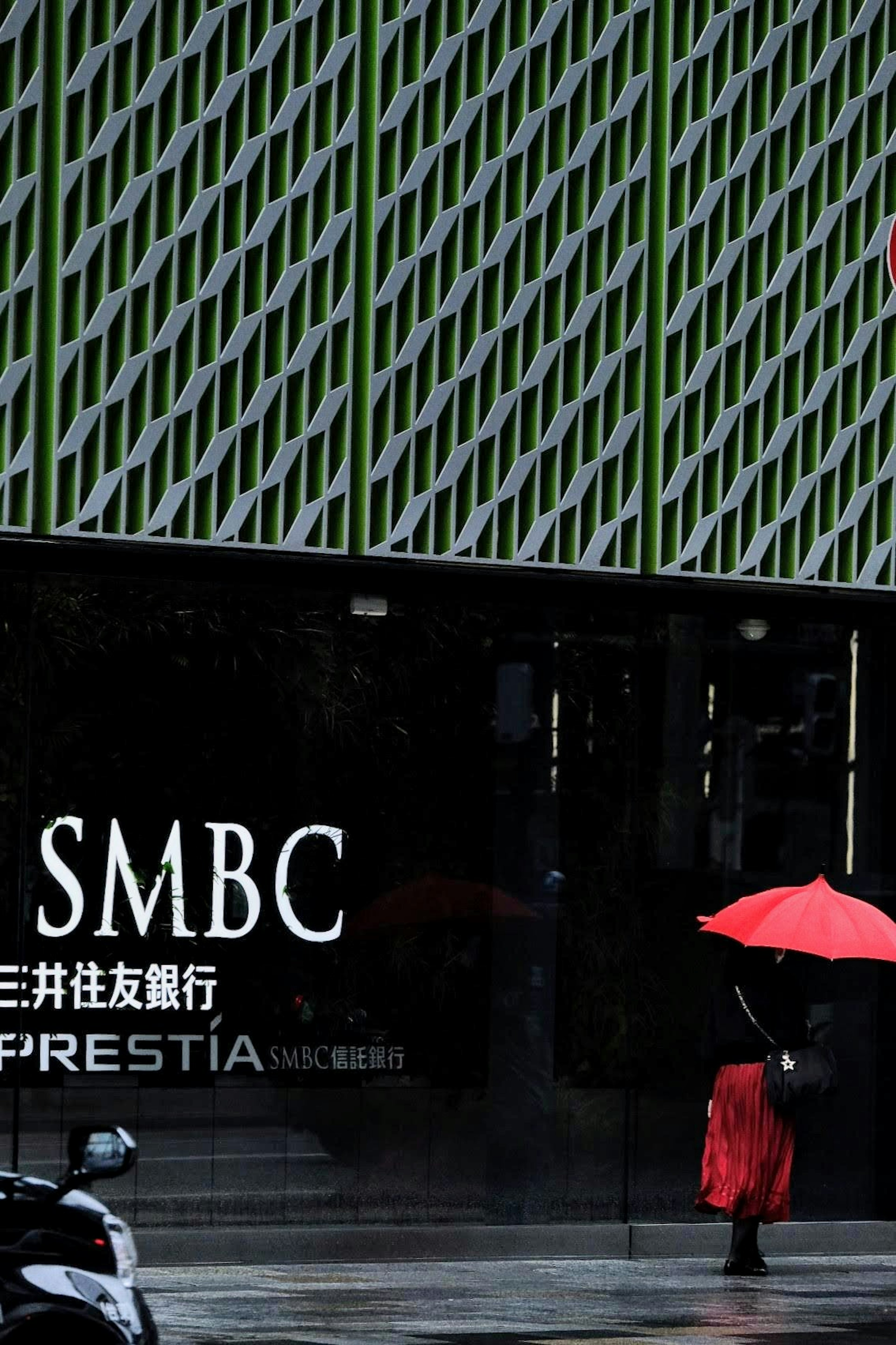 A person holding a red umbrella stands in front of SMBC bank in an urban setting