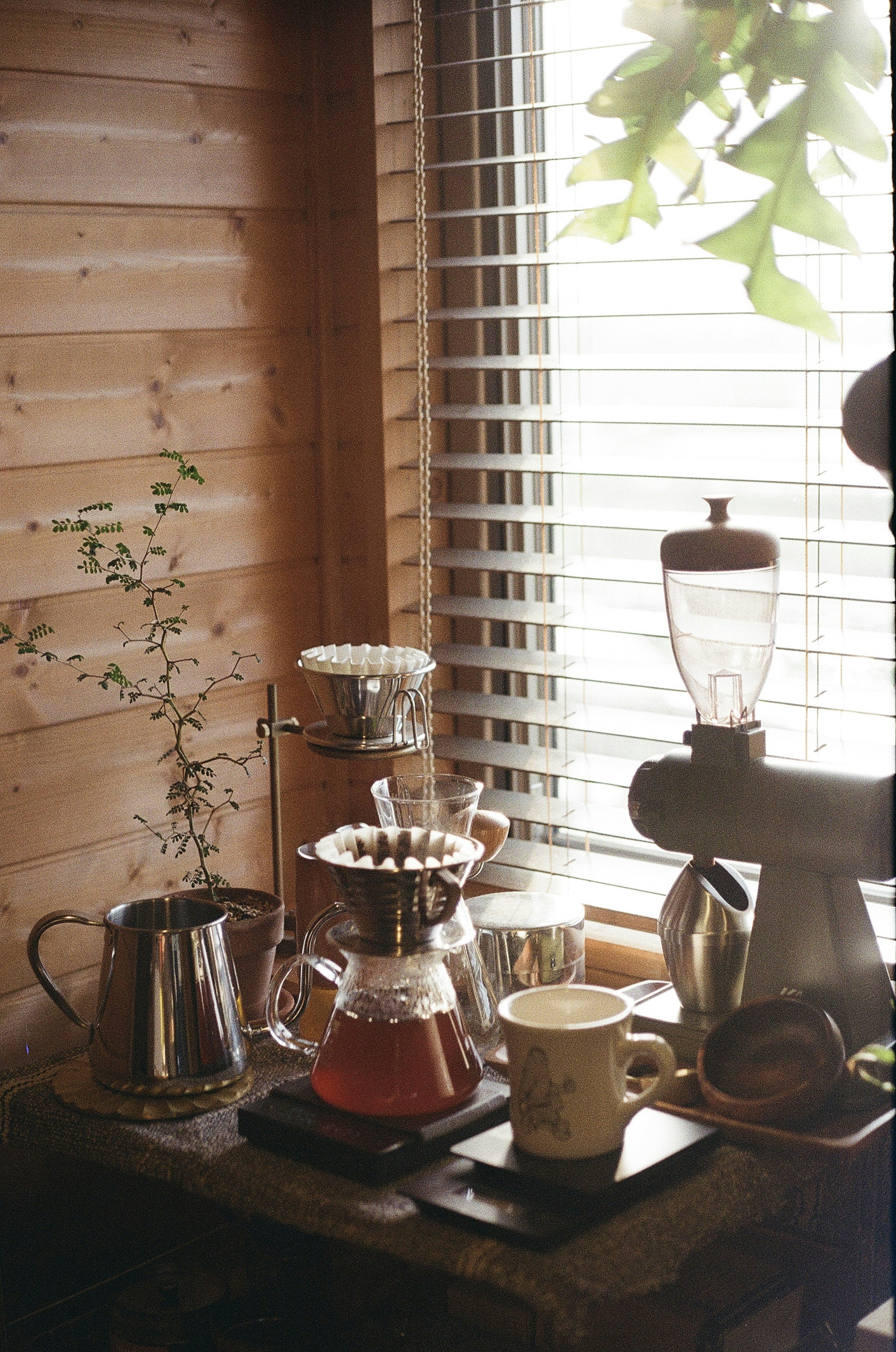 Configuración de preparación de café junto a una ventana con paredes de madera y persianas que presenta varias herramientas de café y una taza