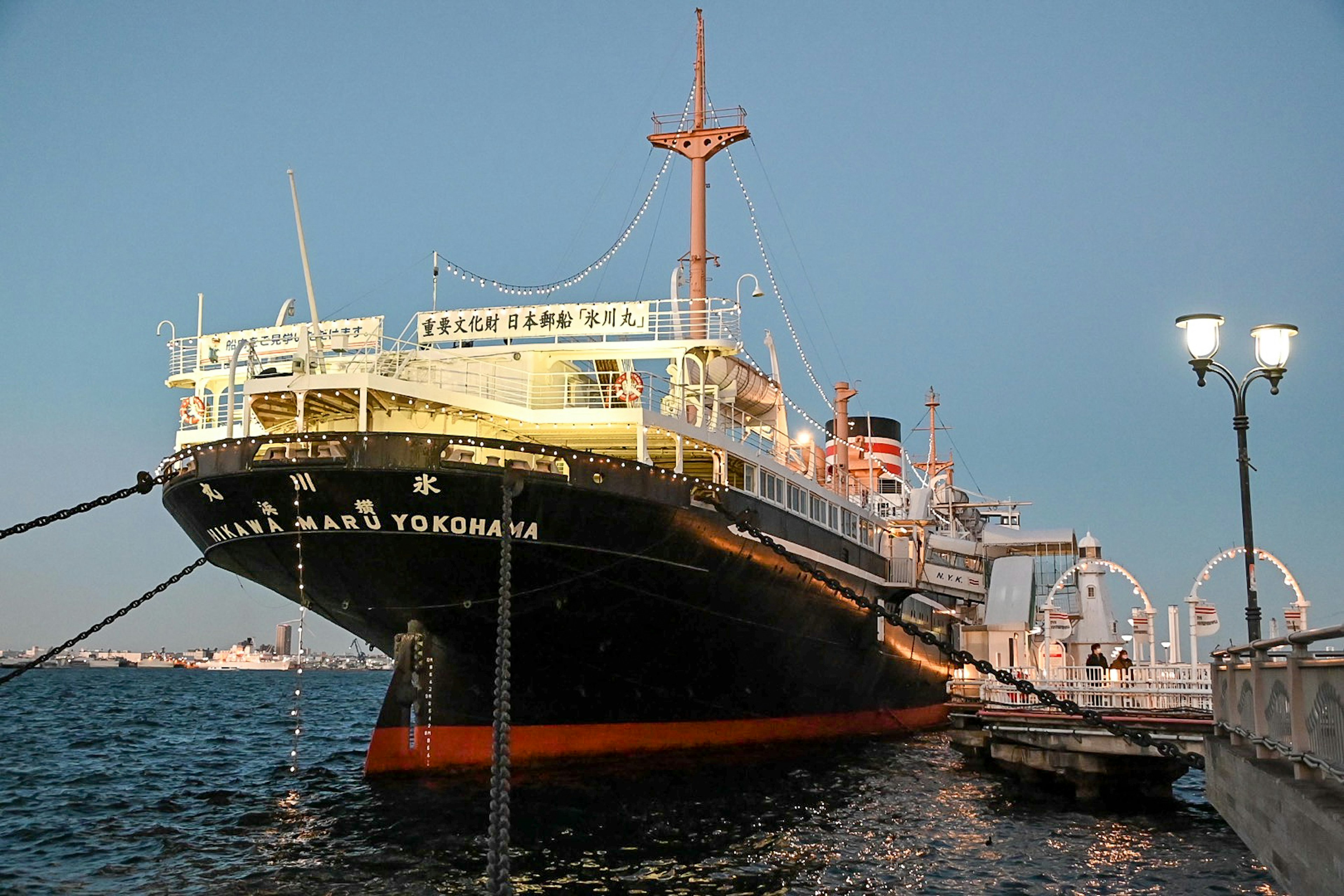 ภาพเรือประวัติศาสตร์ที่จอดอยู่ในน้ำสีฟ้า เรือชื่อ 'Nippon Maru' มีลำตัวสีดำและขาวที่ถูกไฟส่องสว่าง