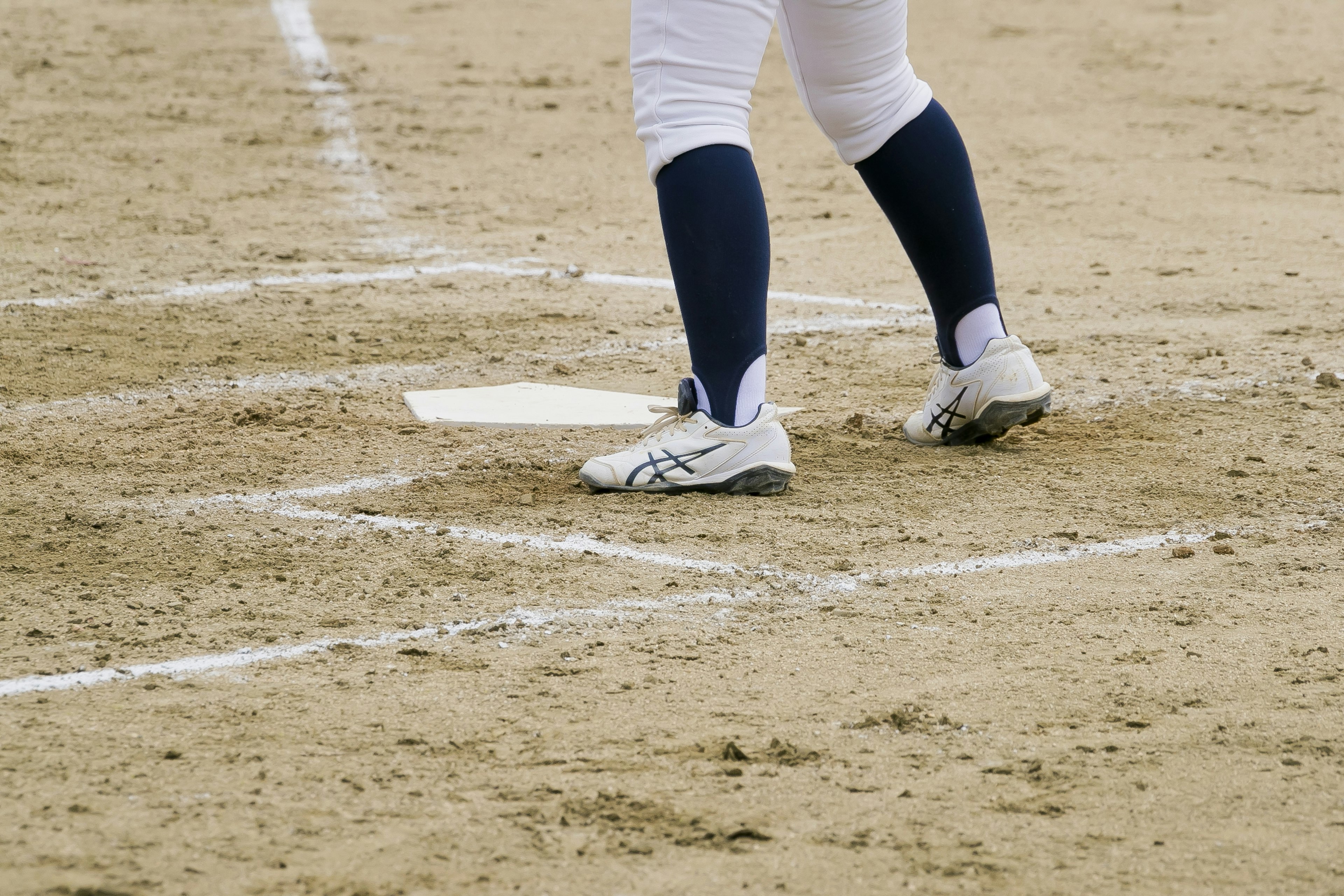 Gambar dekat kaki pemain di lapangan baseball mengenakan sepatu putih dan kaus kaki biru navy