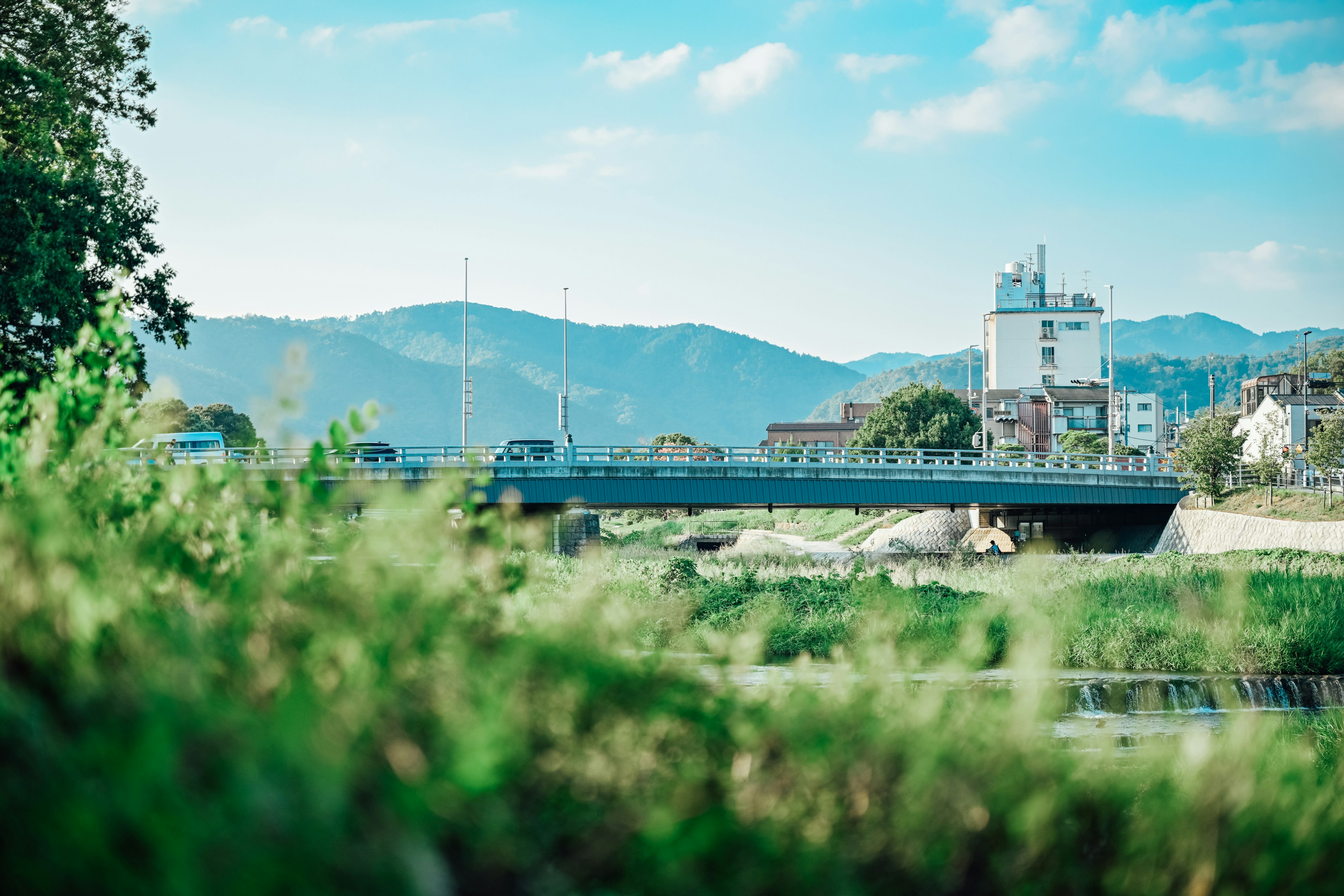 Pemandangan indah sungai dan jembatan di bawah langit biru dengan rumput hijau dan pegunungan di latar belakang