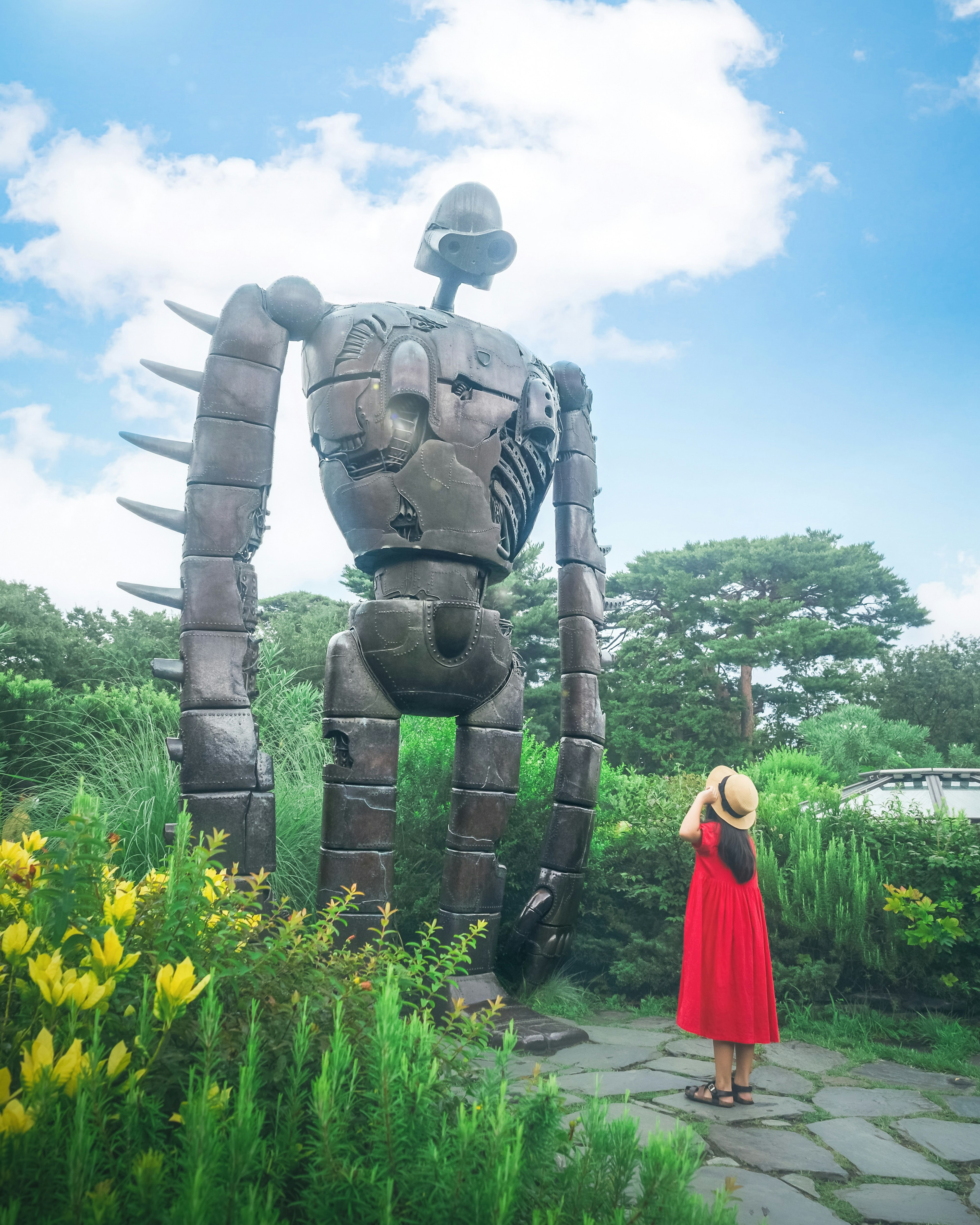 A woman in a red dress stands before a large robot statue surrounded by greenery