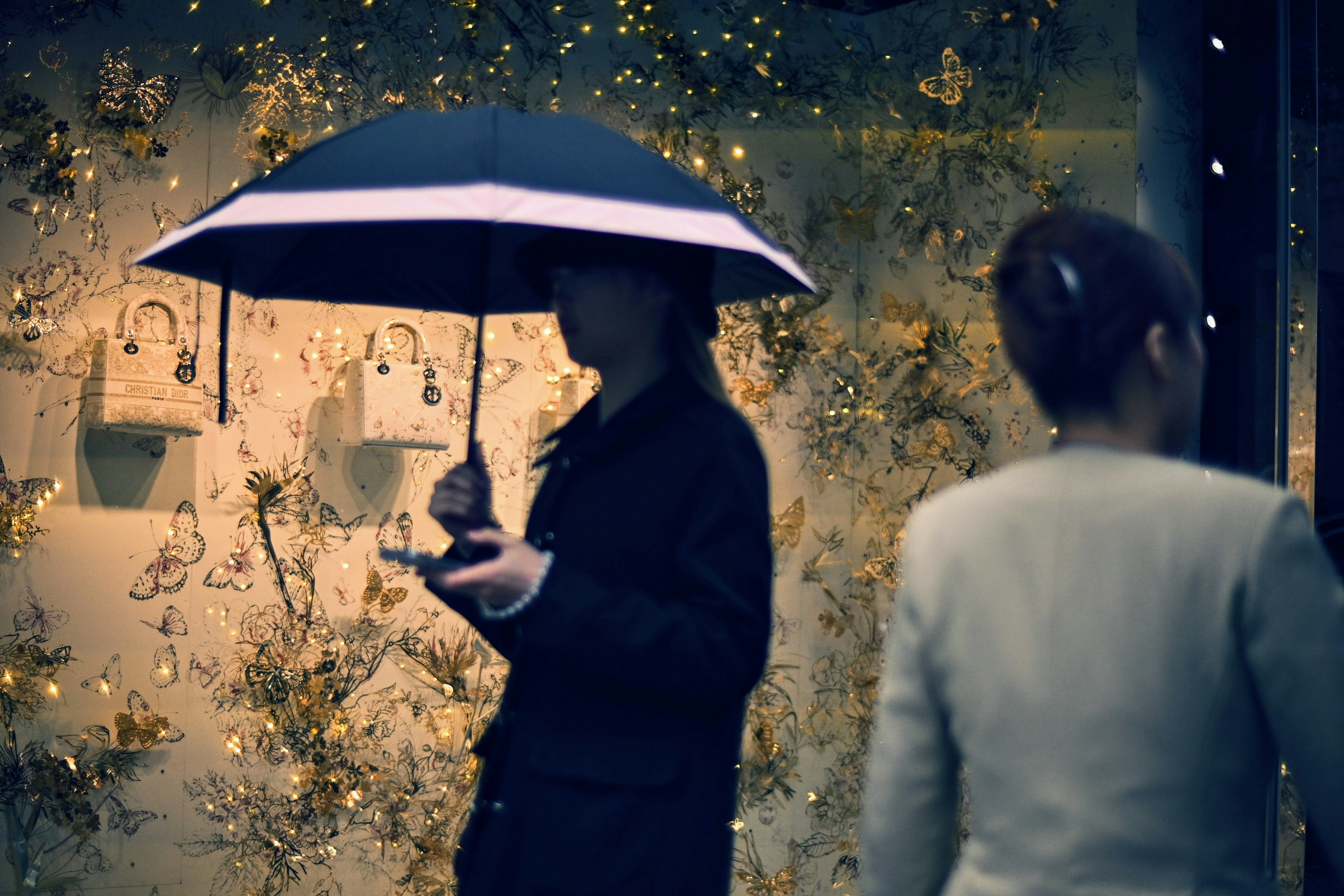 A person holding an umbrella stands in front of a shop window adorned with flowers and lights