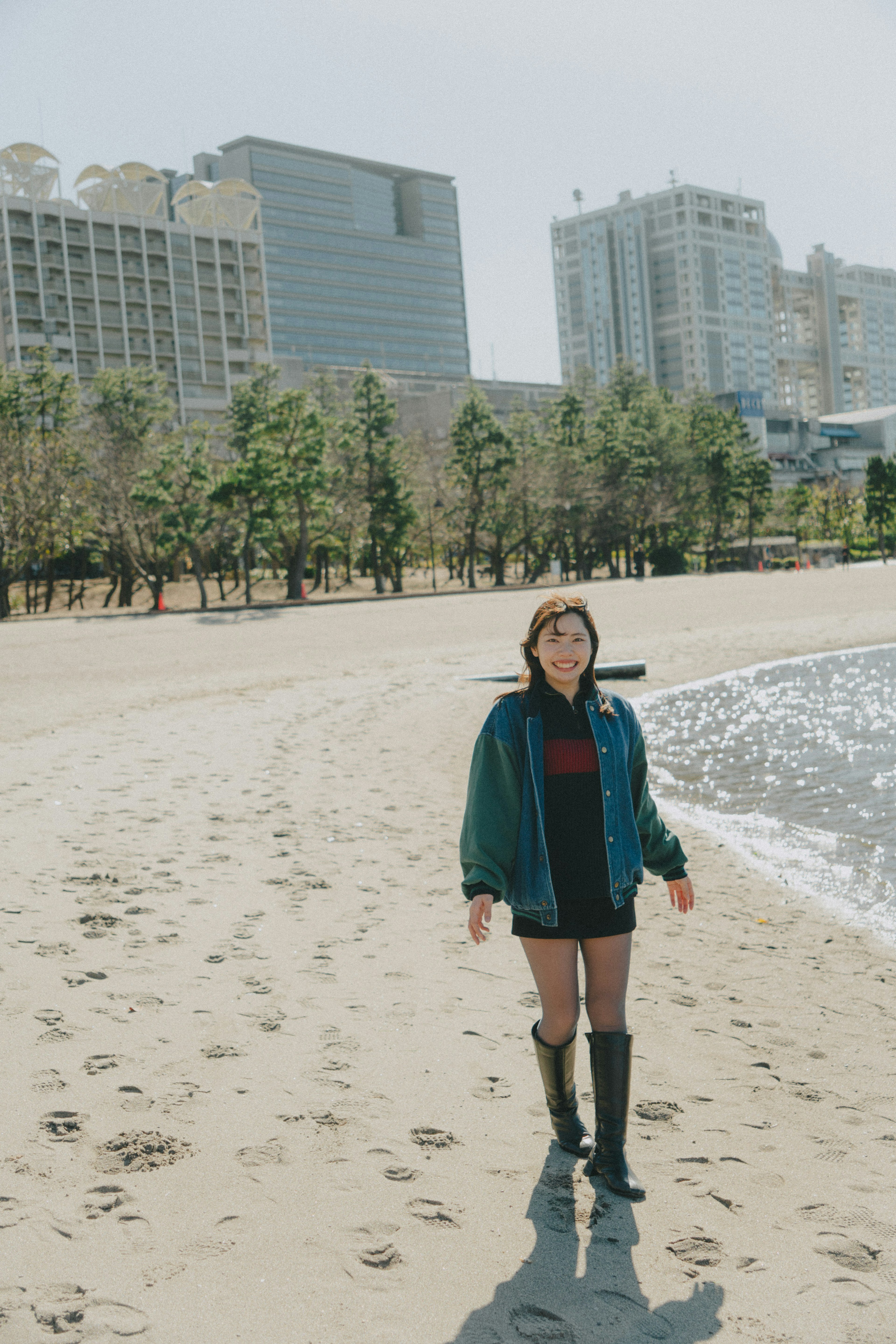 Femme se tenant sur la plage portant une veste bleue et un short noir entourée de grands immeubles et d'arbres