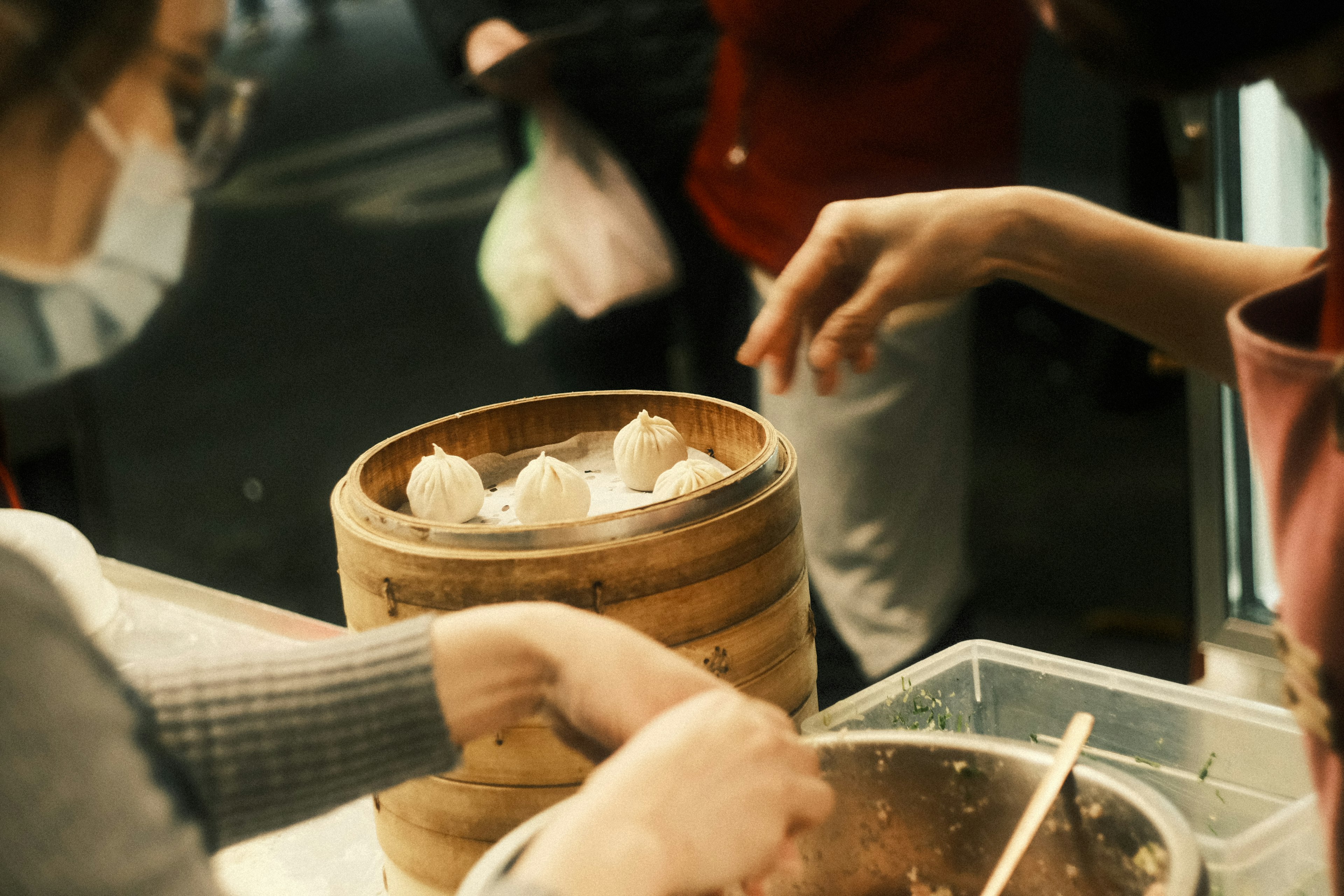 Hands reaching for dumplings from a bamboo steamer