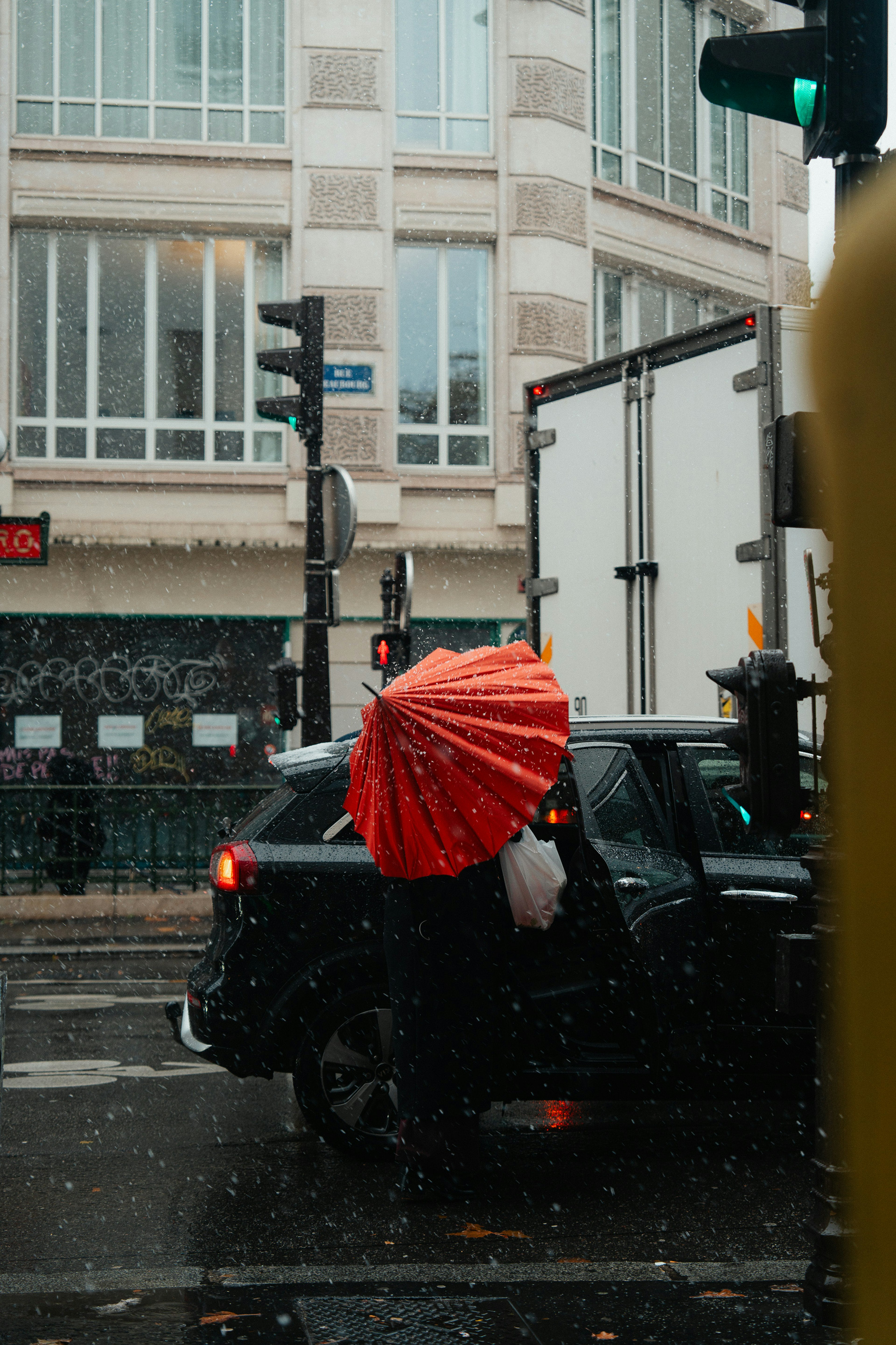 雨の中、赤い傘をさした人物が黒い車の隣にいる風景