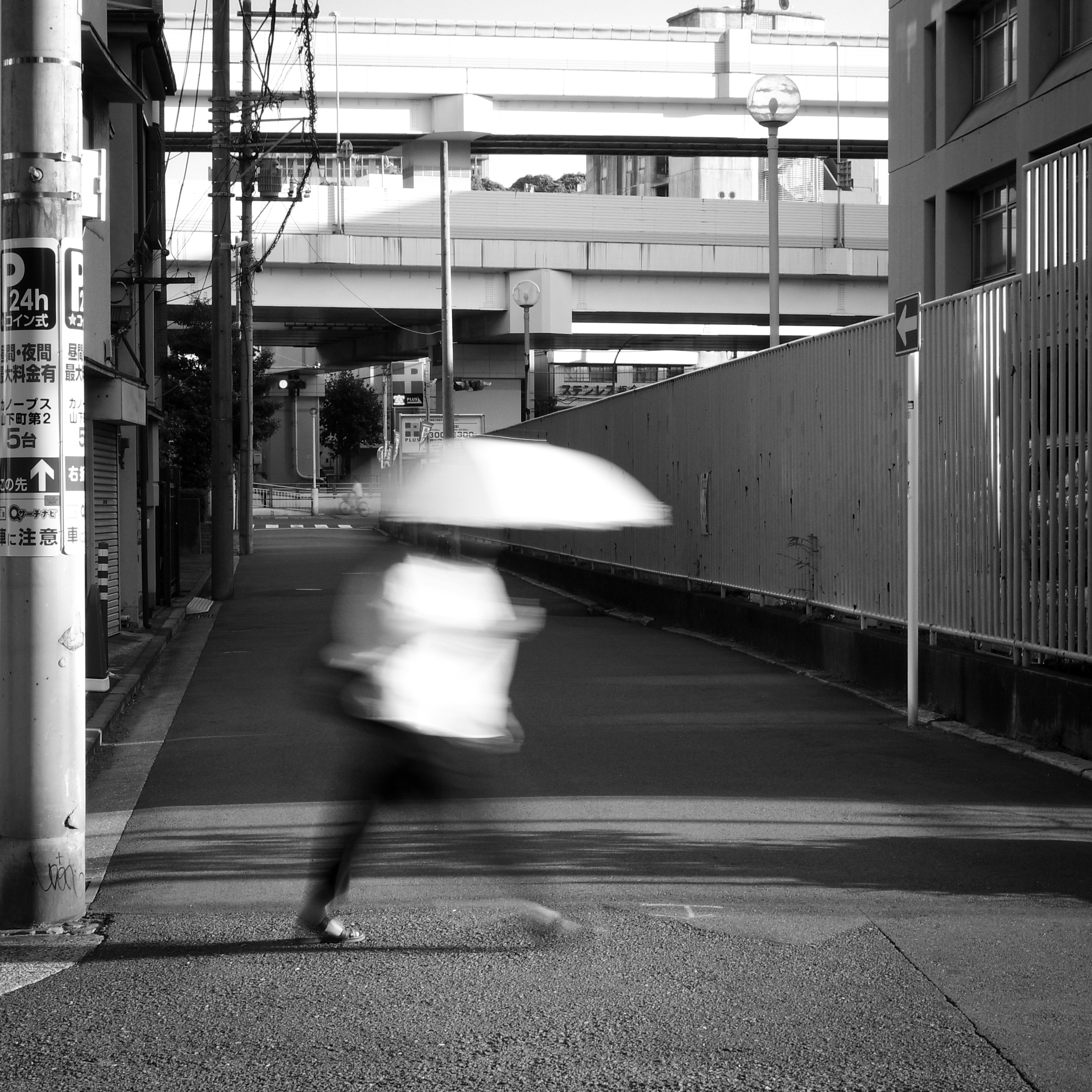 Eine verschwommene Figur, die mit einem Regenschirm in einer Schwarz-Weiß-Straßenszene mit einer erhöhten Straße geht