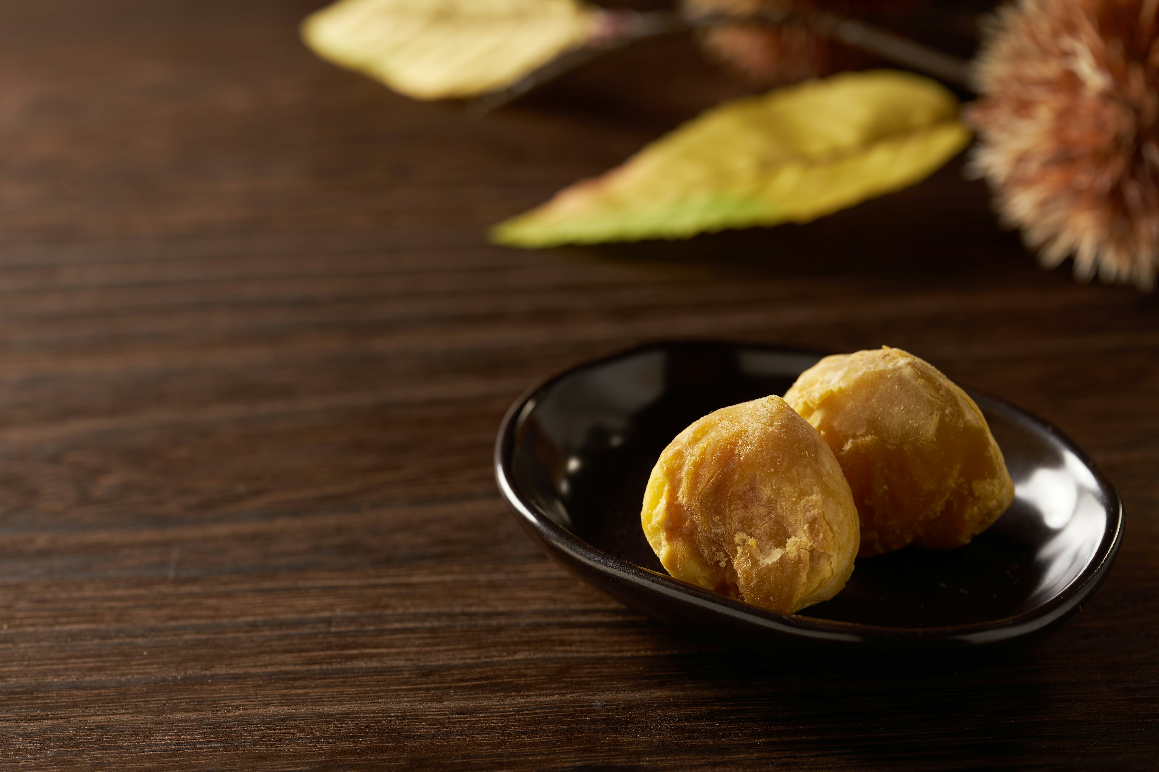 Two chestnut-like food items on a black plate with autumn leaves in the background