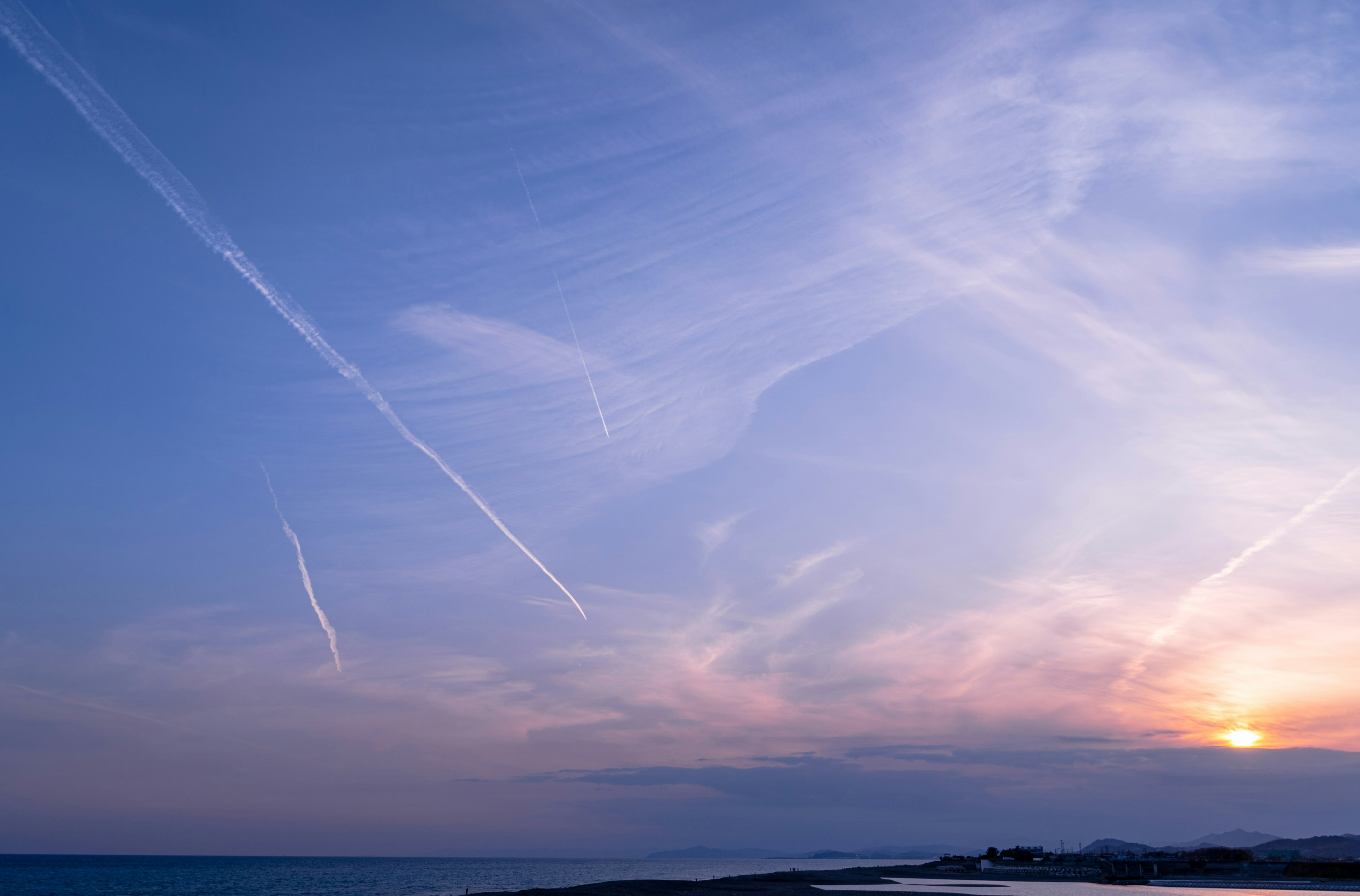 青い空に雲と飛行機雲が広がり夕日が沈む海の風景