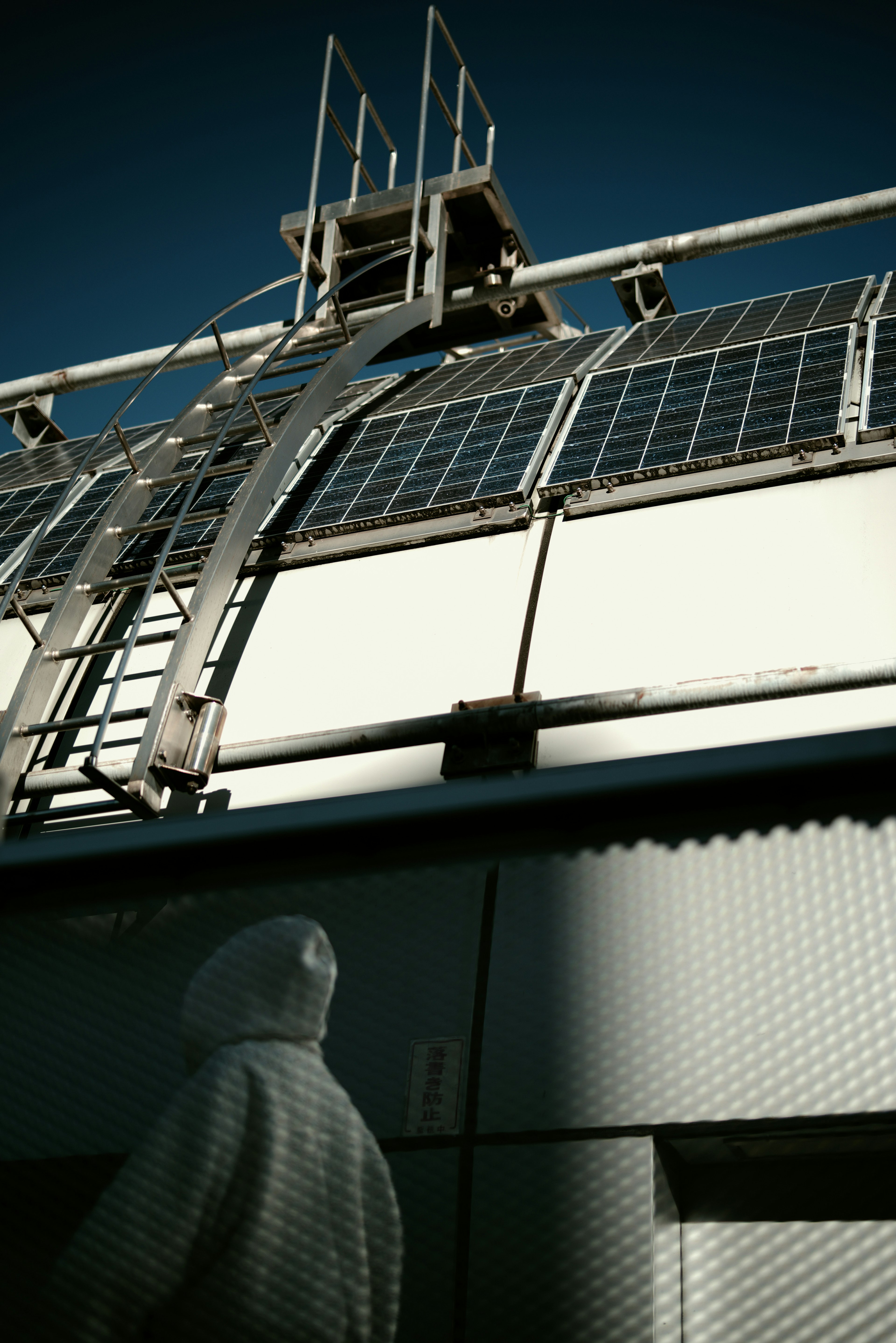 A person in white work attire stands beside a building featuring a metal ladder and grid roof in the background