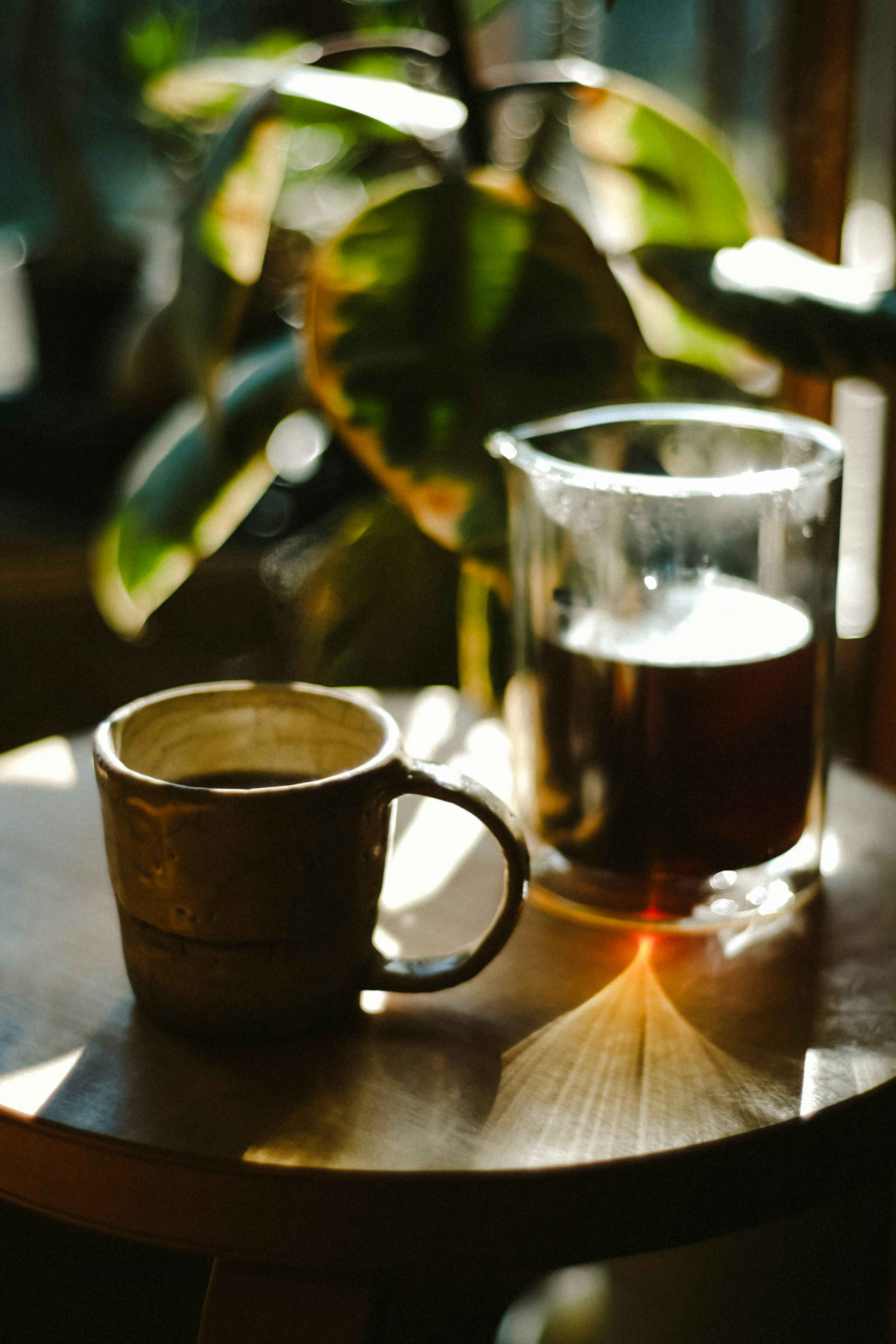 Cangkir kopi dan wadah kaca di atas meja kayu dengan tanaman di latar belakang