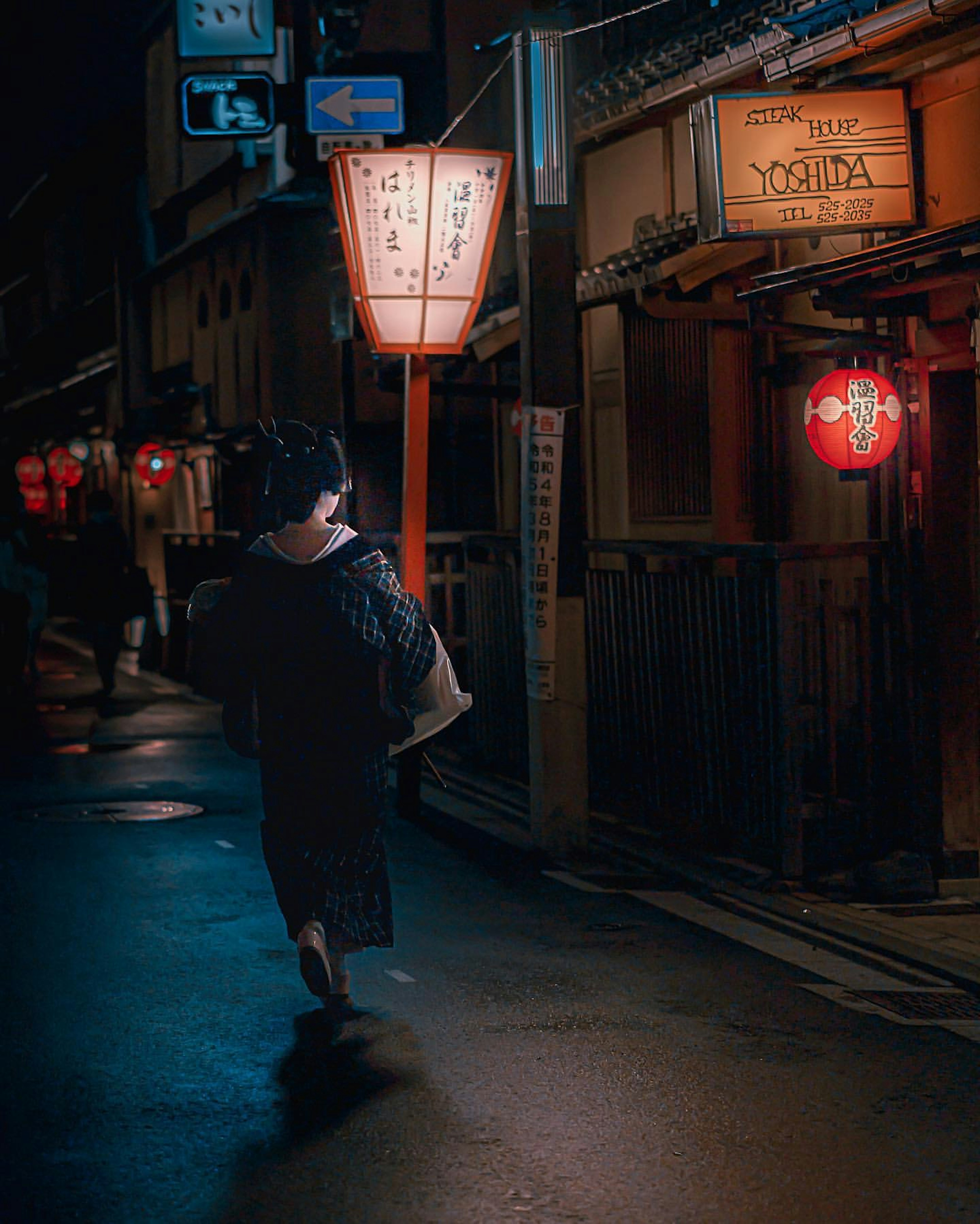 Eine Frau im Kimono, die durch eine nächtliche Straße geht, beleuchtet von roten Laternen