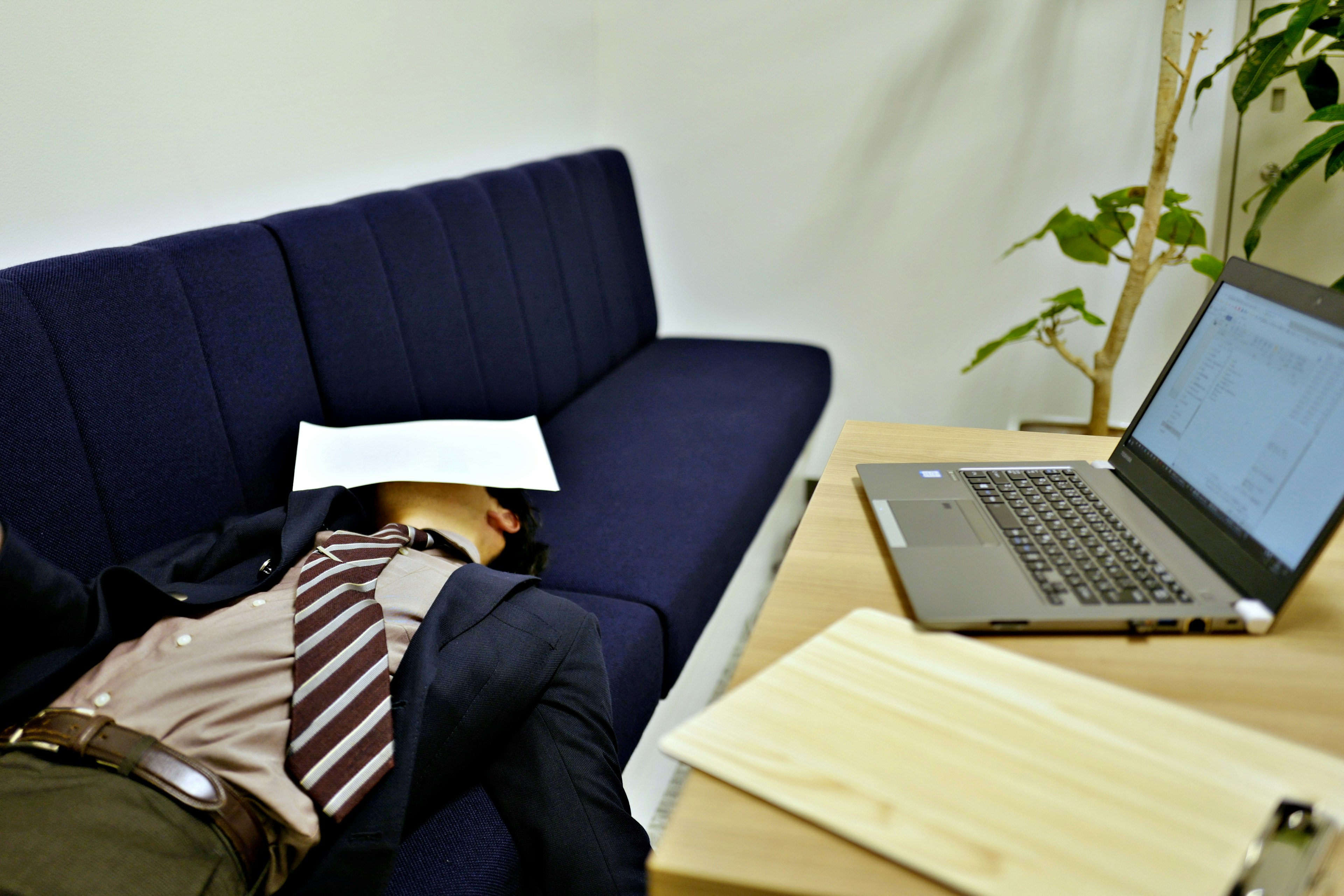 Geschäftsmann schläft auf einem blauen Sofa mit Laptop und Dokumenten in der Nähe