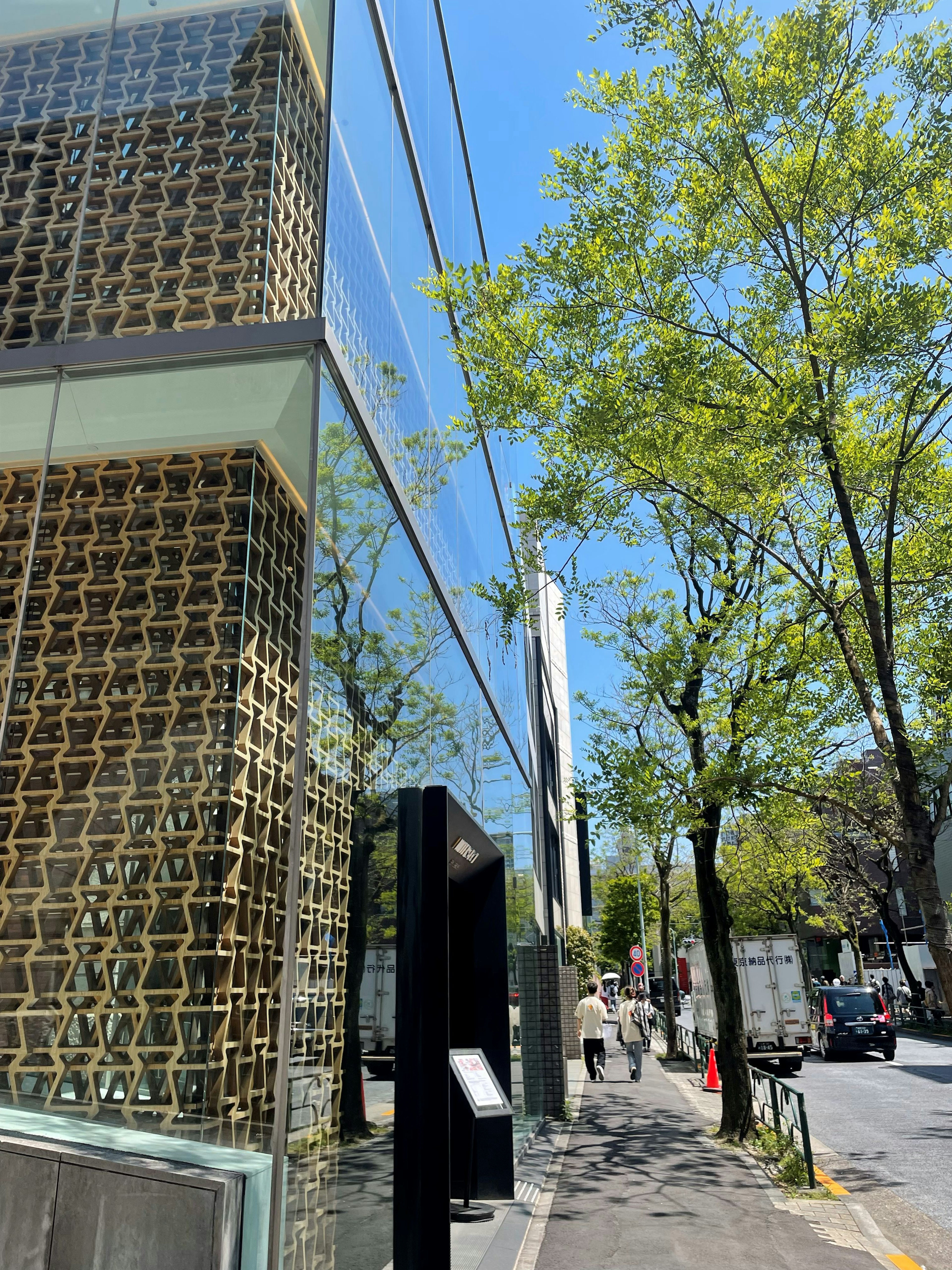 Immagine che mostra la facciata di un edificio moderno rivolto verso la strada con un muro a design intrecciato e alberi verdi sotto un cielo blu