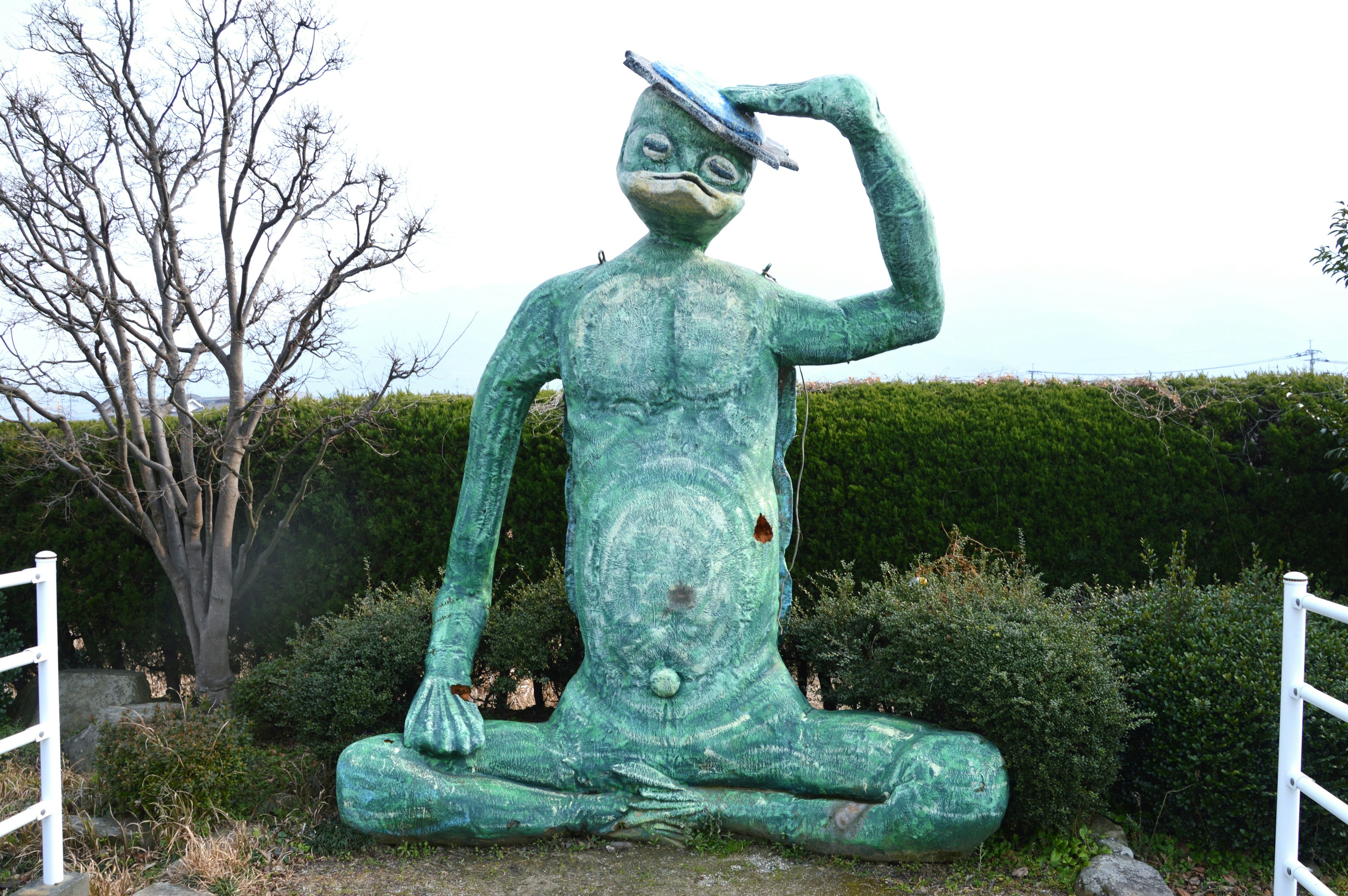 Large green sculpture of a seated figure with a hat on its head surrounded by greenery