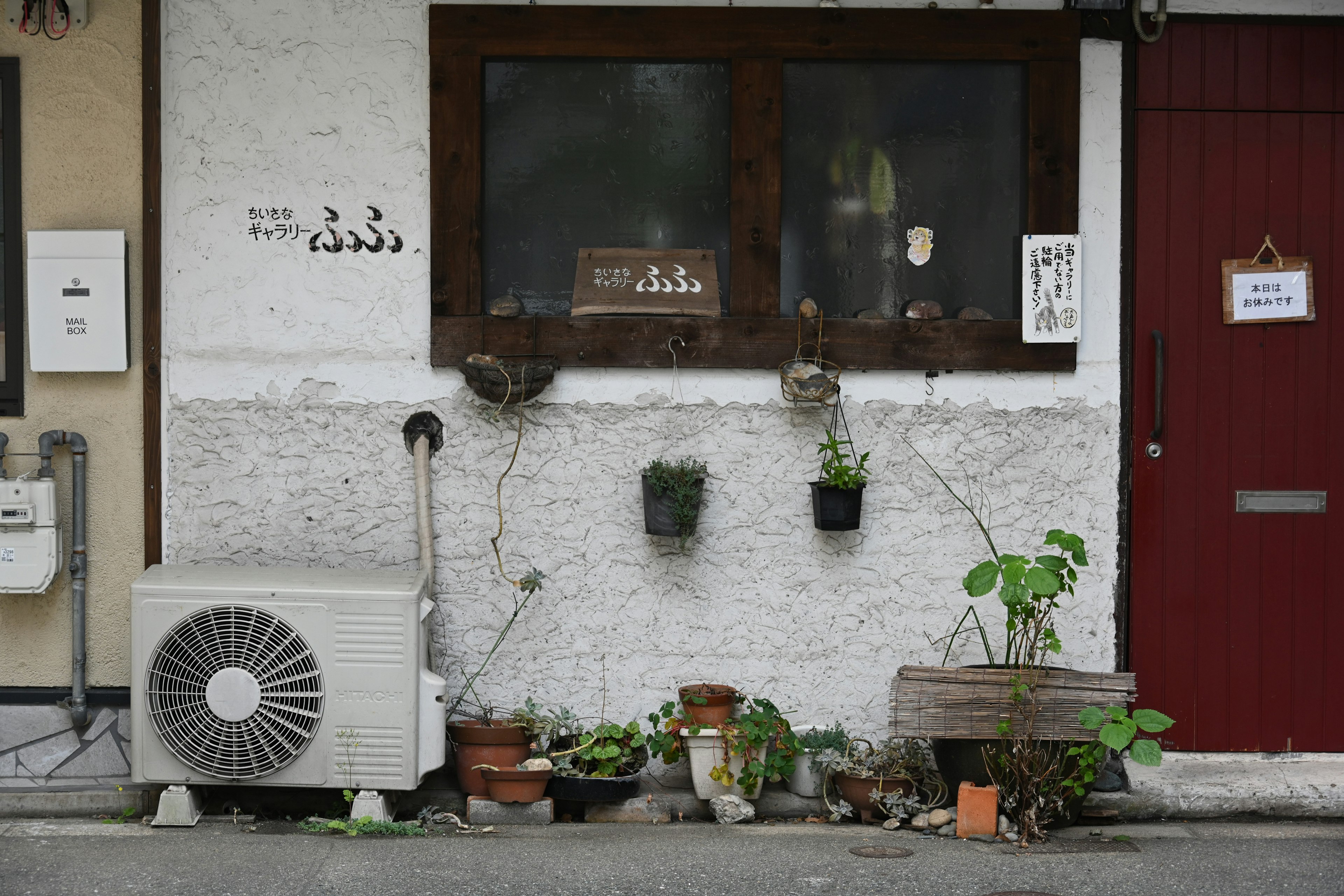 Extérieur d'un magasin avec des murs blancs et une porte rouge comportant des plantes sur la fenêtre et une unité de climatisation