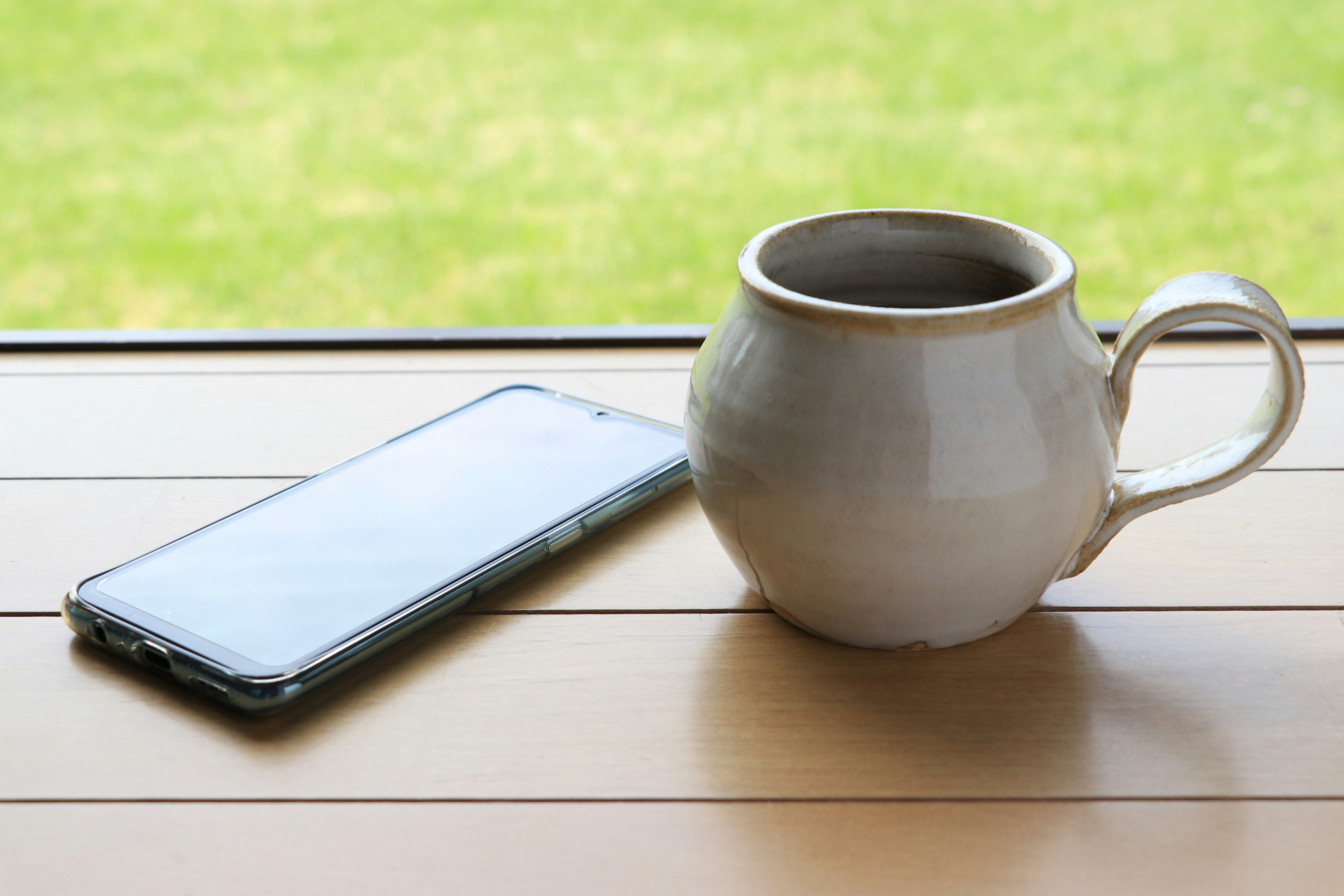 Una tazza da caffè bianca e uno smartphone posati su una superficie di legno vicino alla finestra