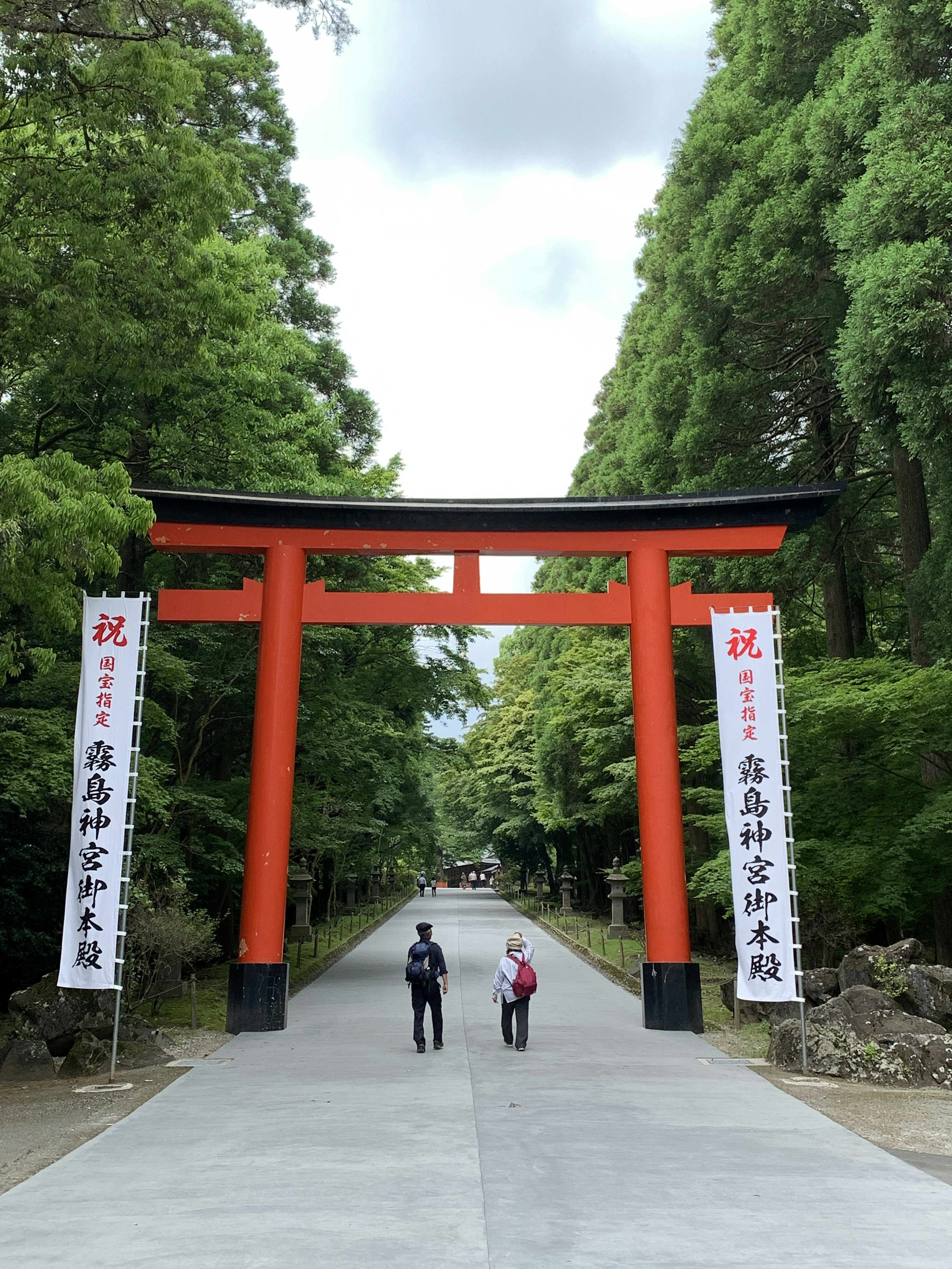 Dua orang berjalan di bawah gerbang torii merah dikelilingi oleh pepohonan hijau