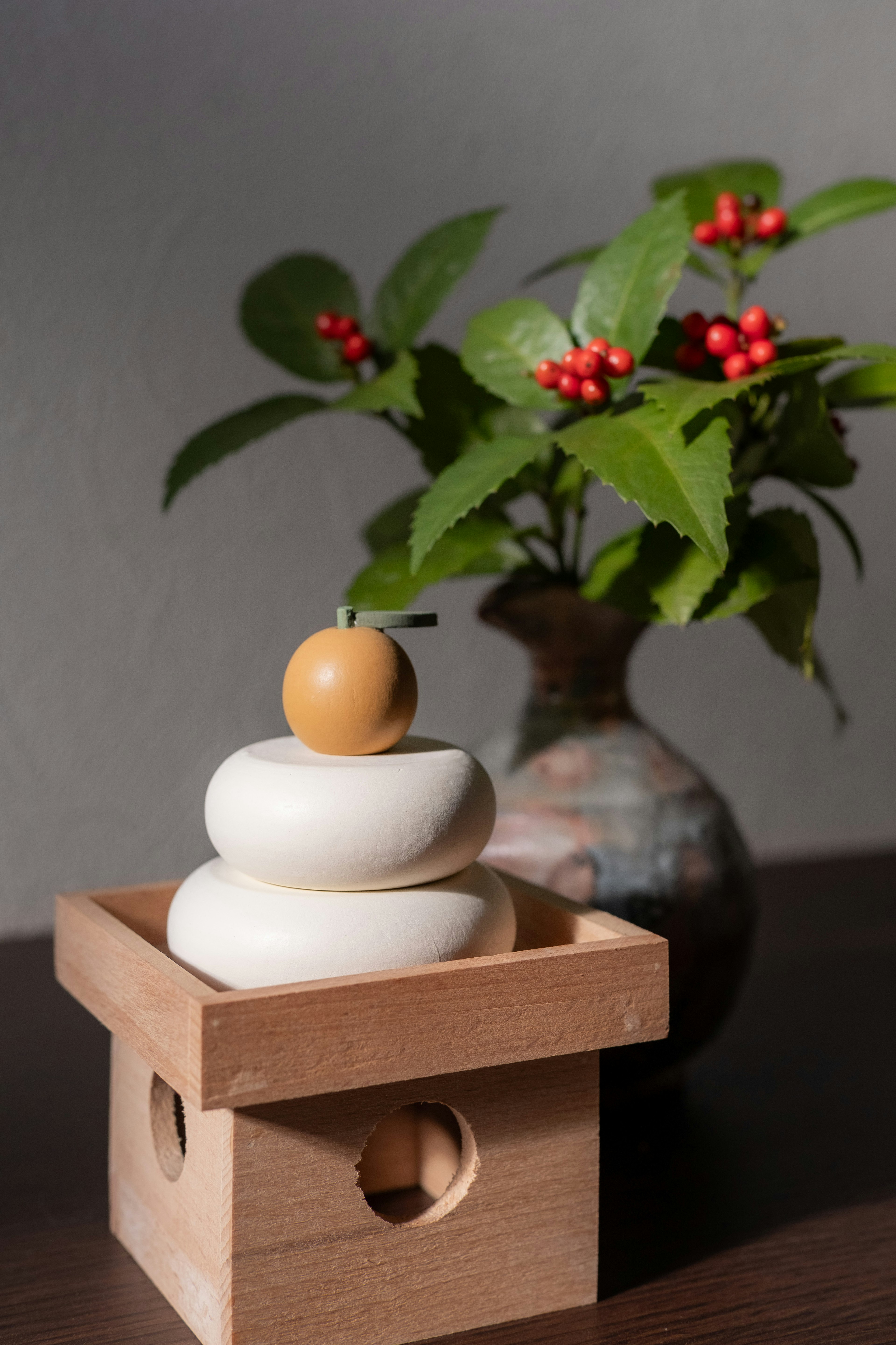 Image of stacked white circular objects topped with an orange sphere in a wooden box with a vase of red berries