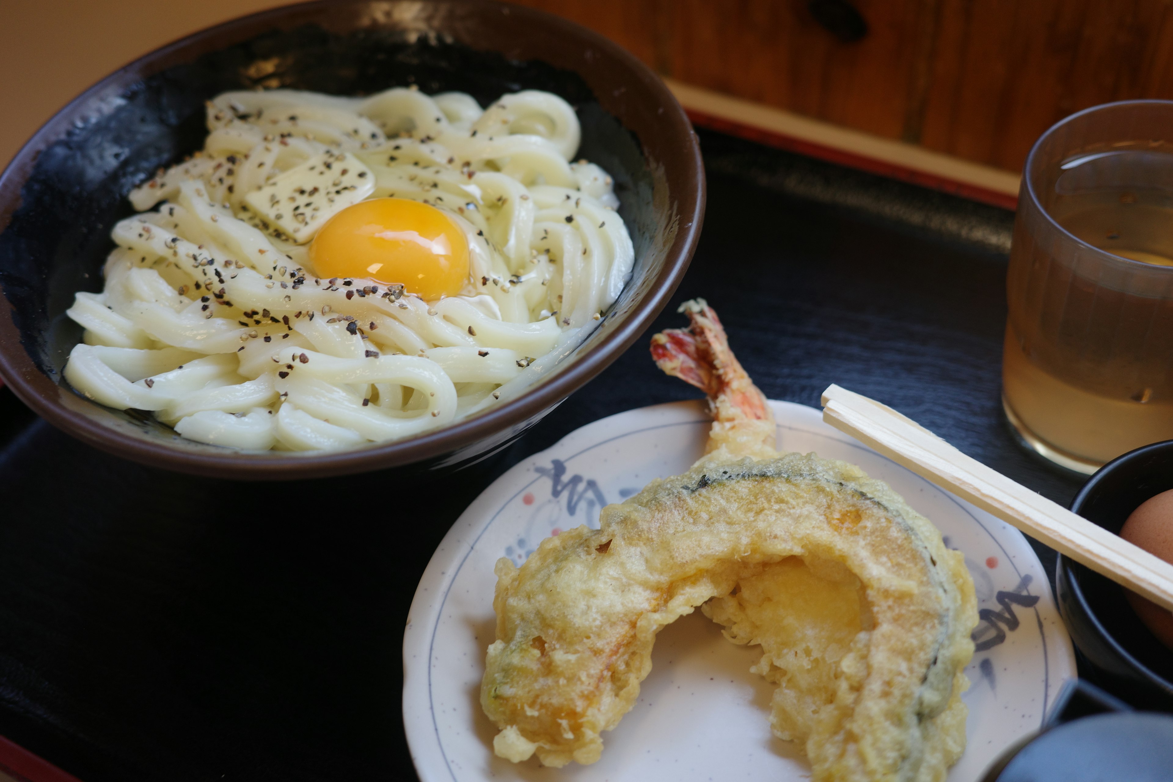 Semangkuk mie udon dengan telur mentah dan piring tempura udang serta sayuran