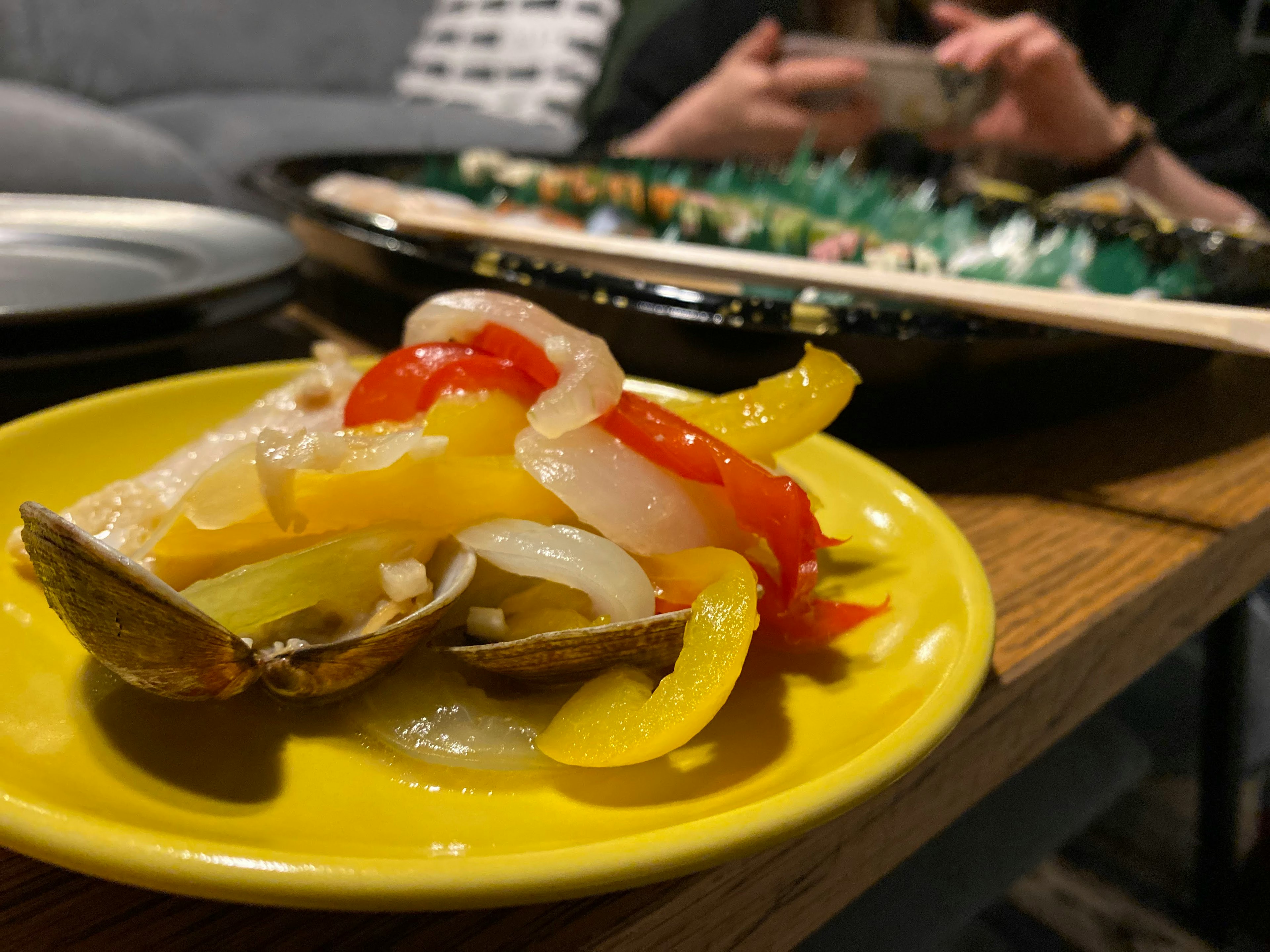 Verduras coloridas y almejas en un plato amarillo con sushi al fondo