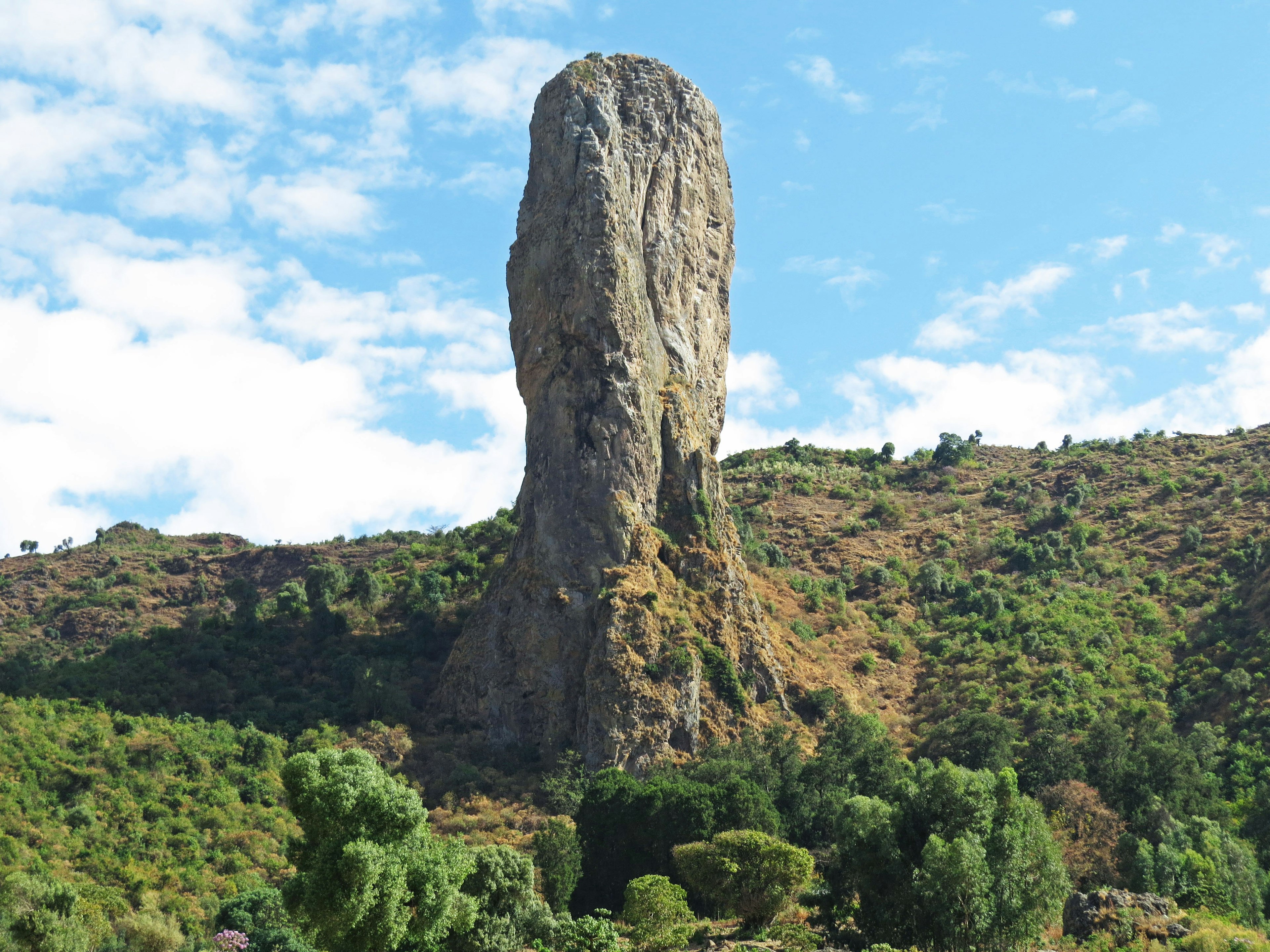 Tall rock formation on green hillside under blue sky