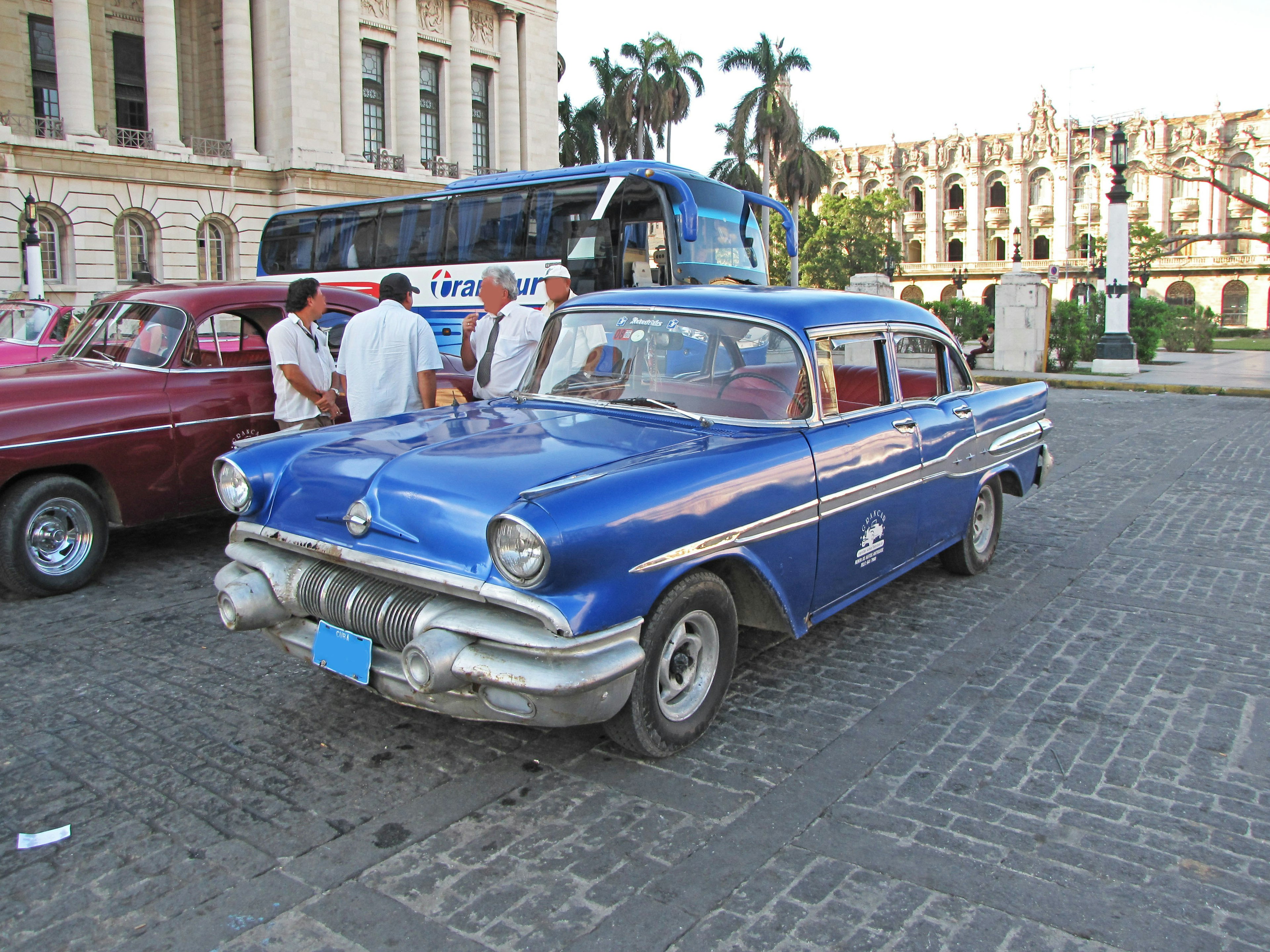 Un coche antiguo azul estacionado en una plaza con personas reunidas alrededor