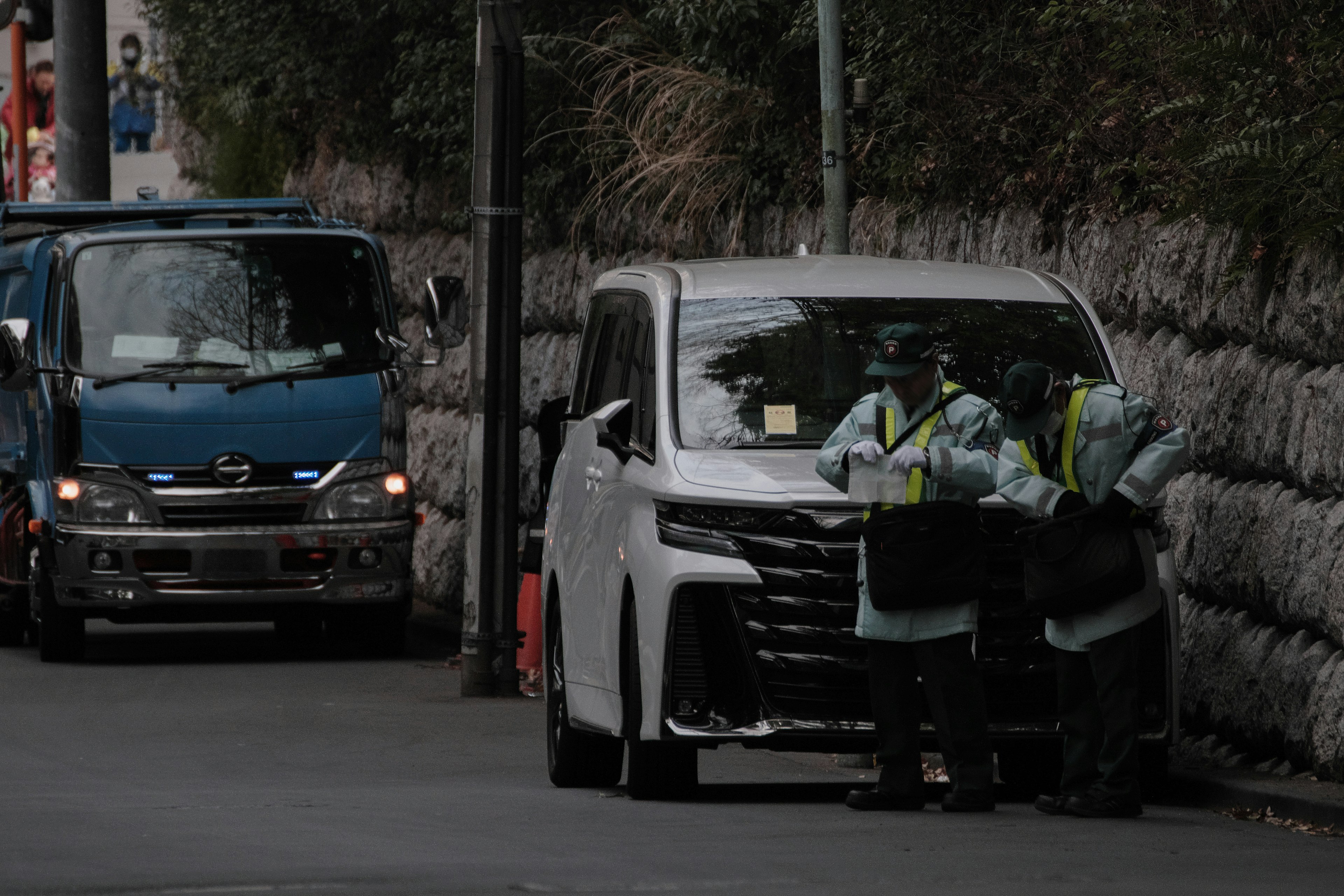 白い車と青いトラックが並ぶ道路で作業する二人の警備員