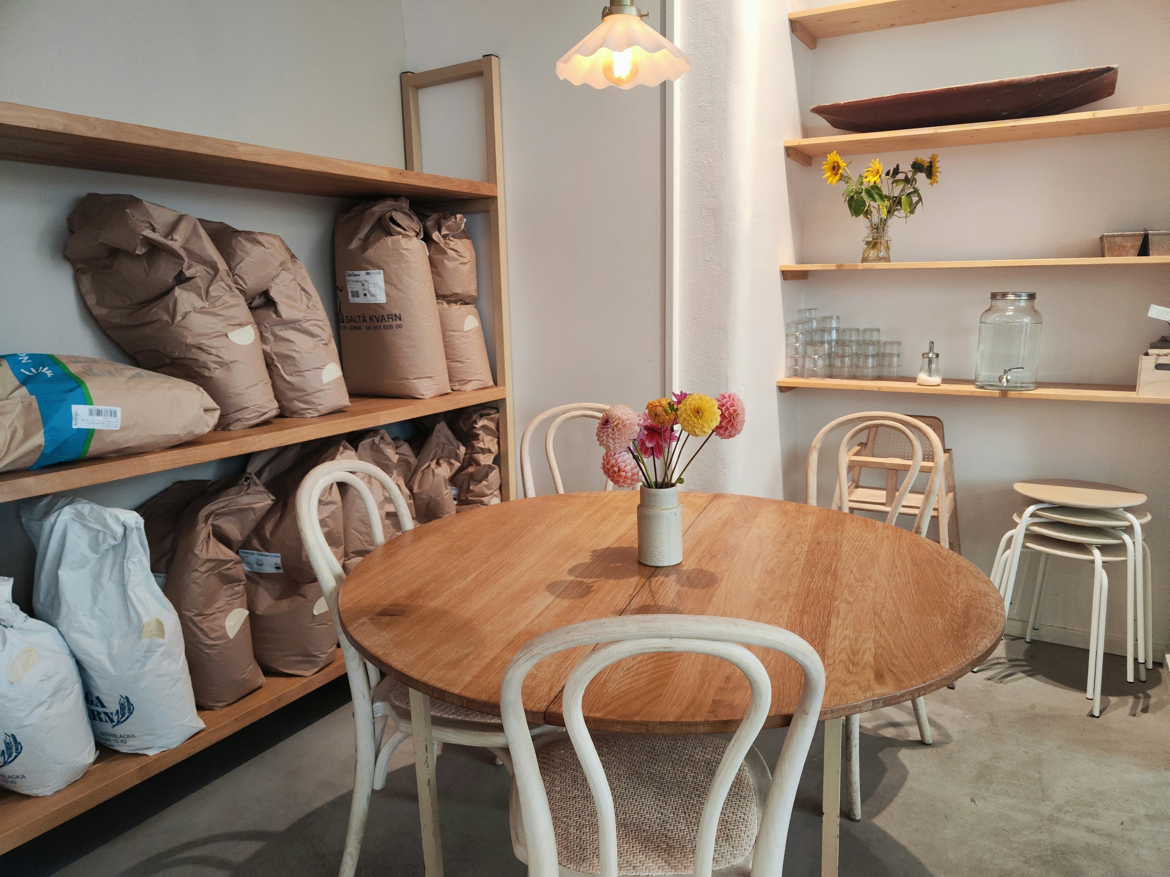 Interior of a café featuring a wooden table and white chairs with bags and a vase on the shelves