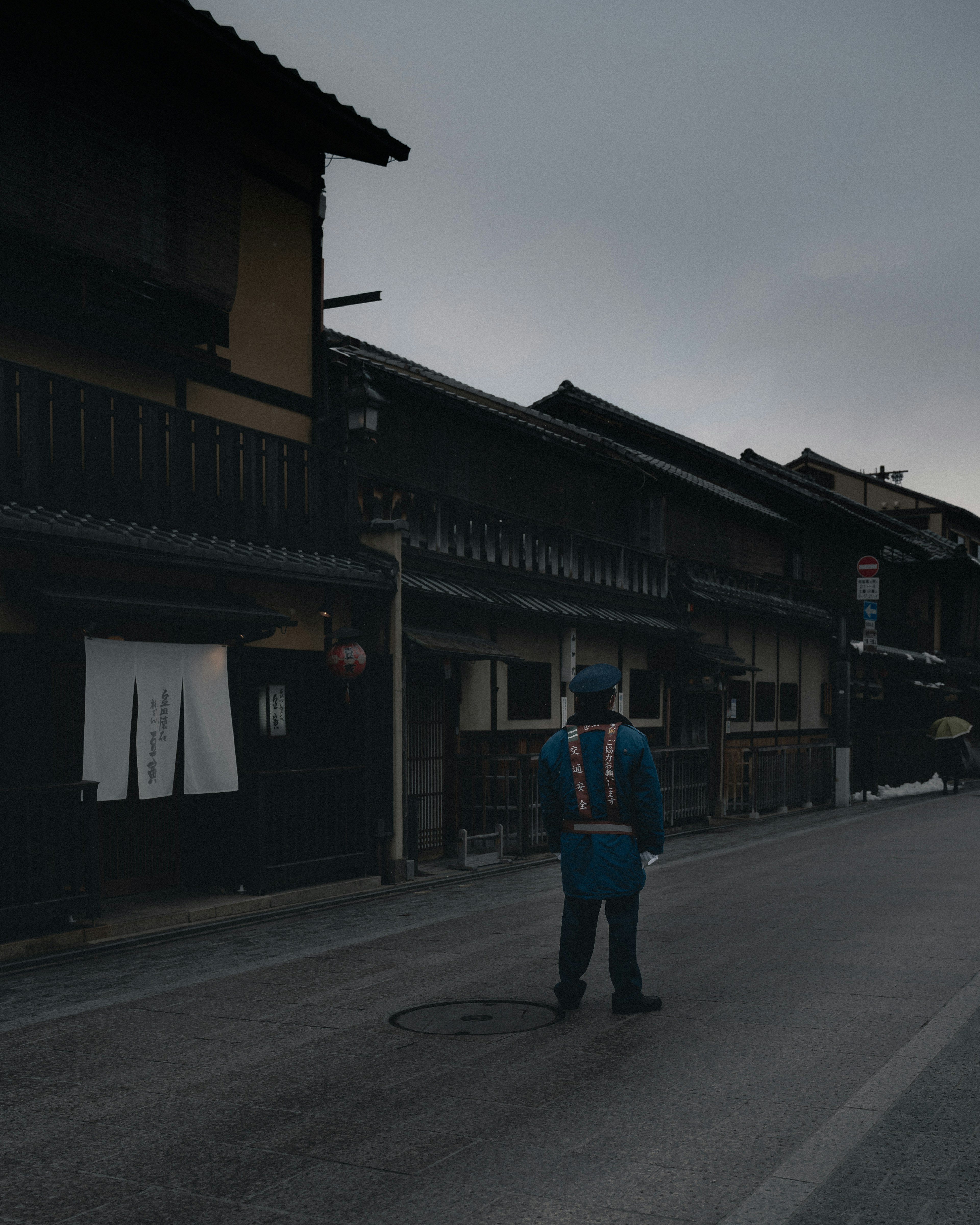 A person in a blue uniform standing on a quiet street lined with traditional buildings