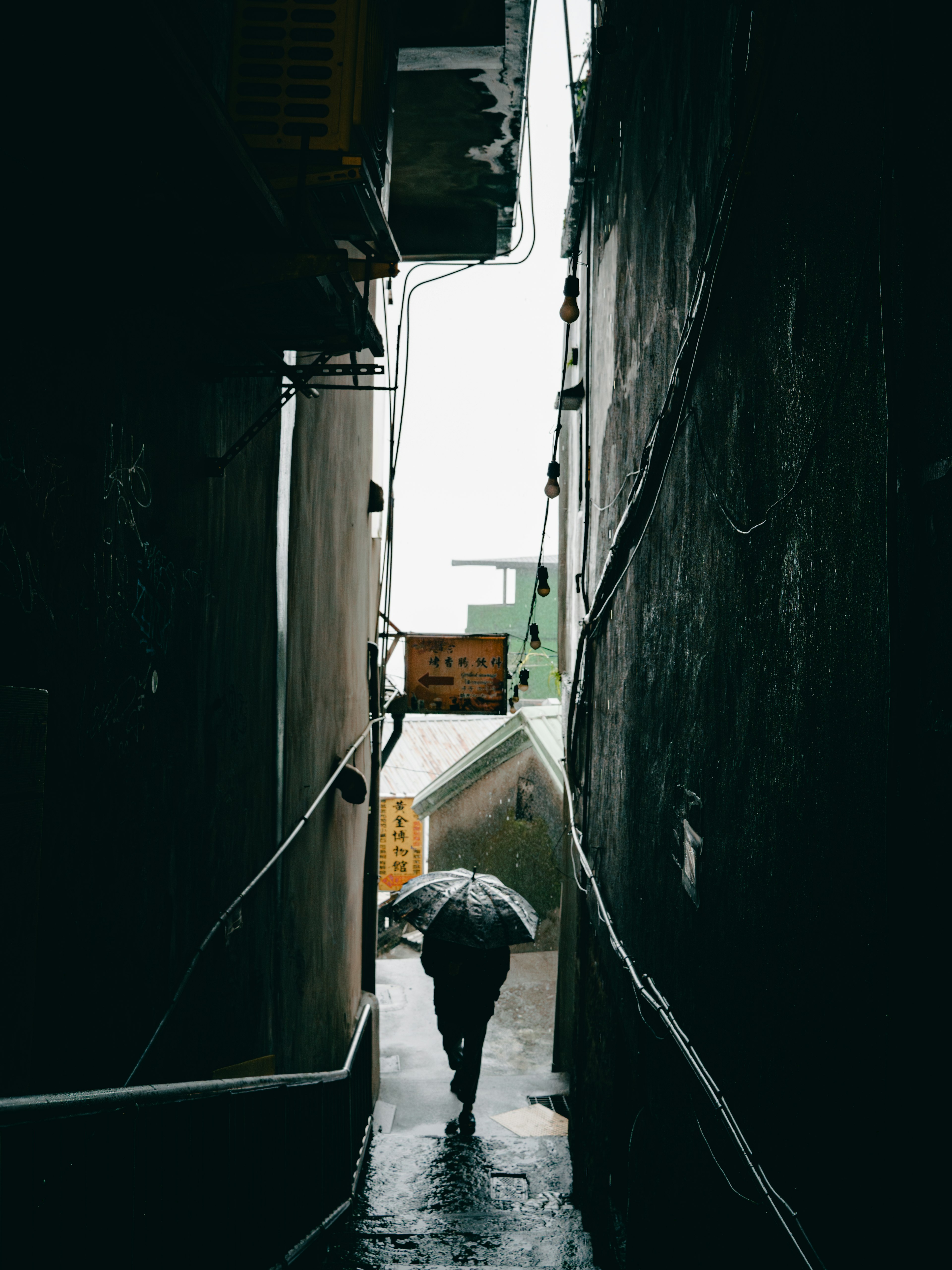 A person walking in a narrow alley holding an umbrella in the rain