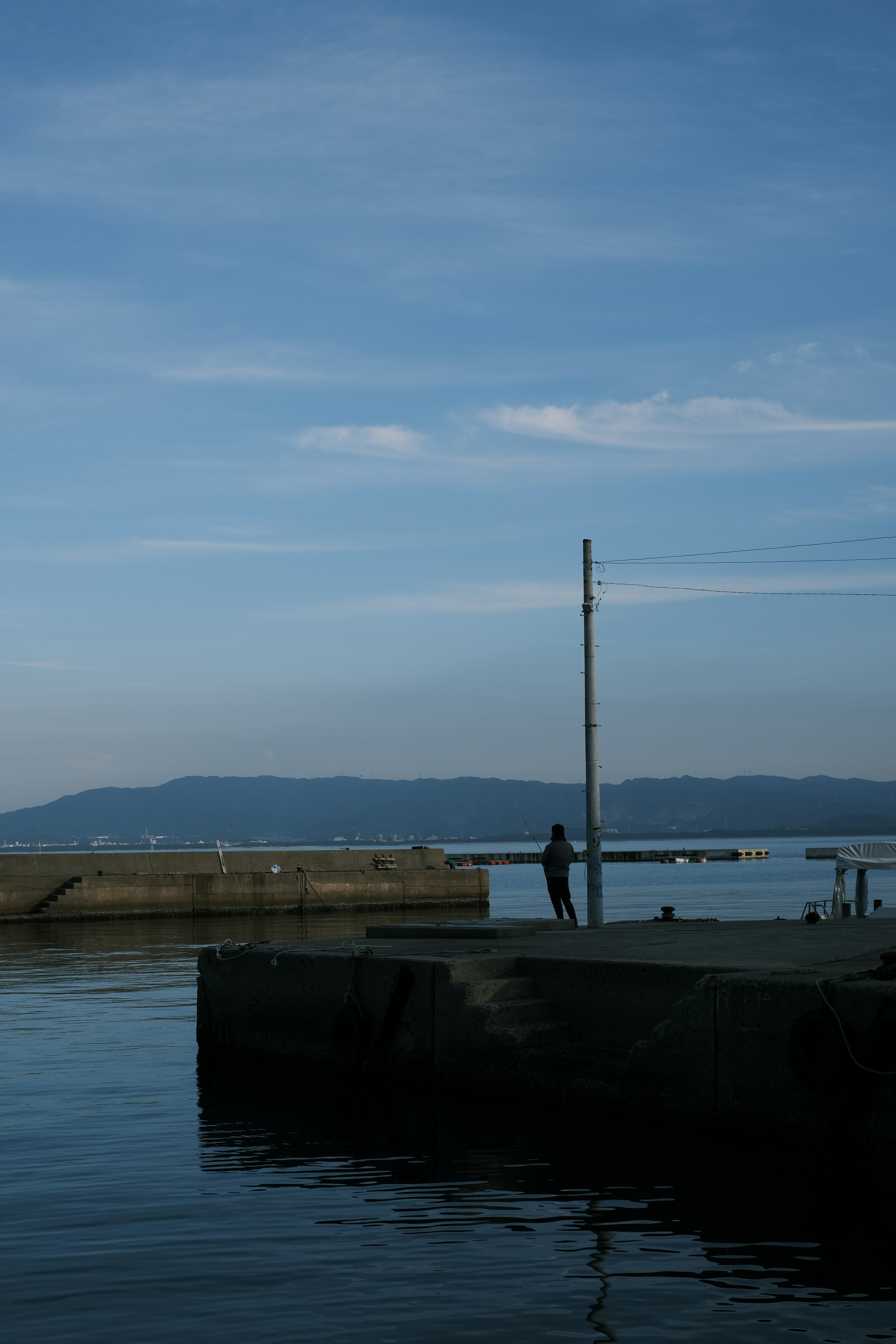 ทิวทัศน์ท่าเรือที่เงียบสงบพร้อมชายคนหนึ่งยืนอยู่บนท่าเรือใต้ท้องฟ้าสีฟ้าและน้ำที่สงบ