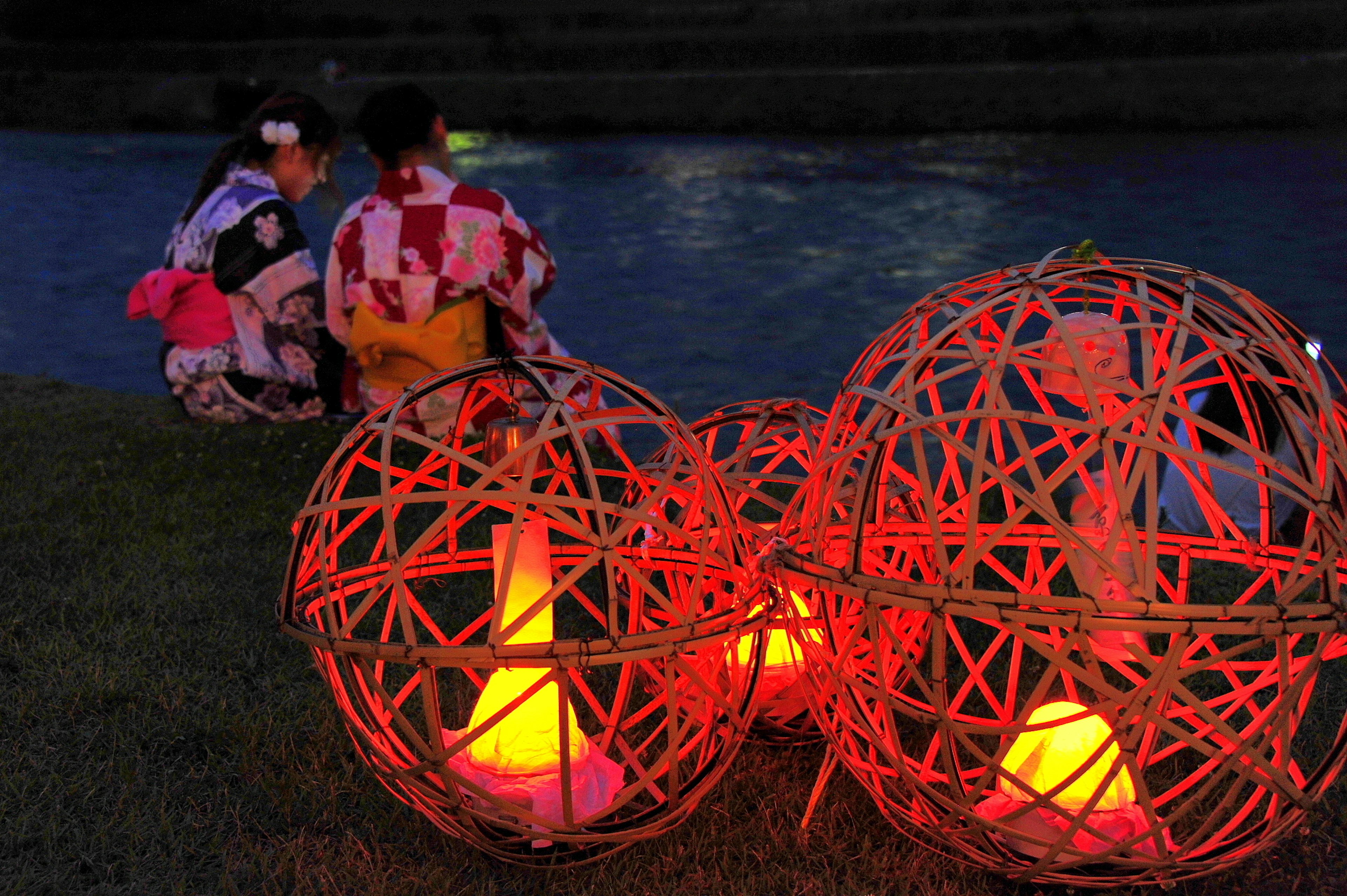 Dos mujeres en kimono sentadas junto a un río por la noche con linternas de bambú rojas brillantes en primer plano