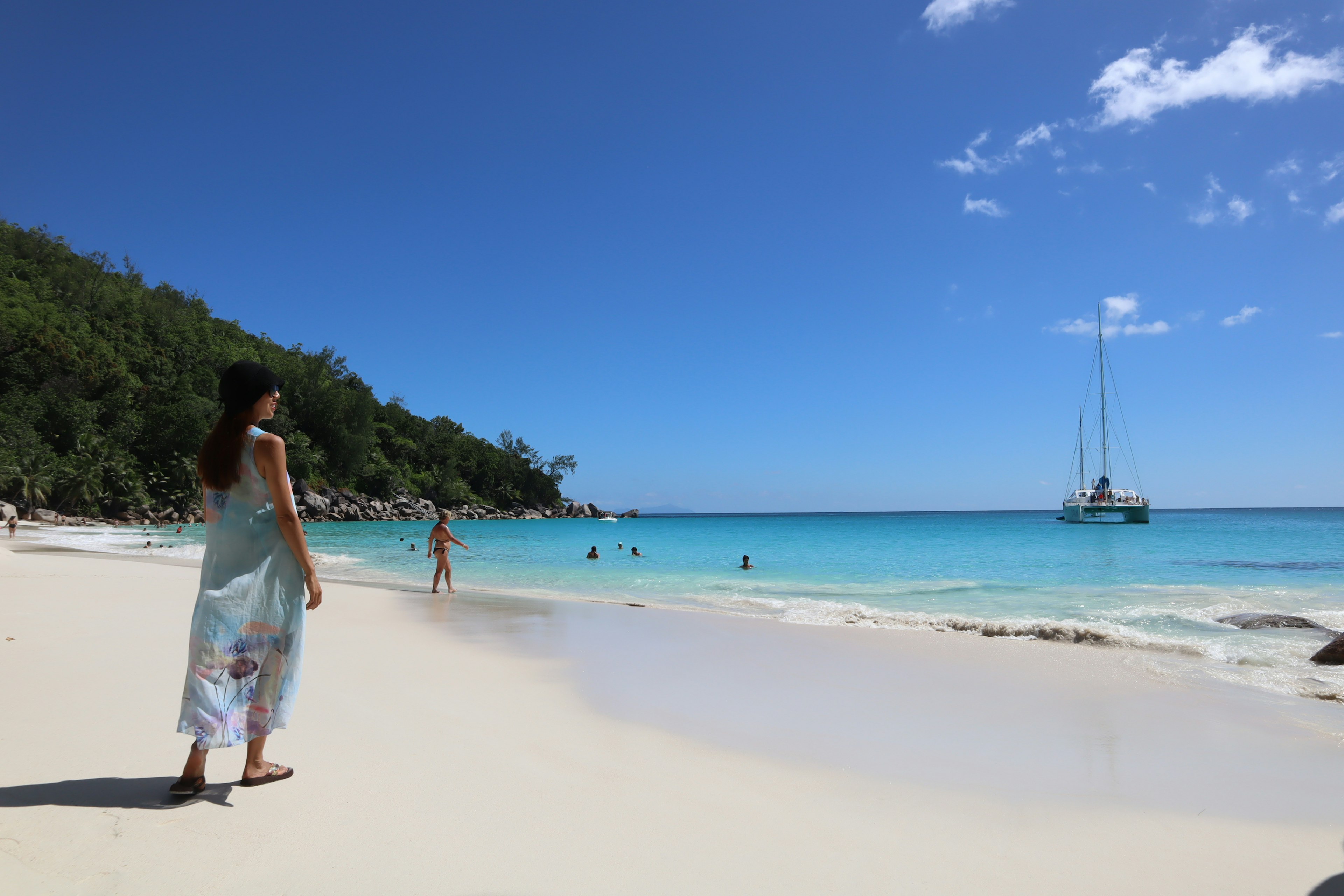 Una mujer caminando en una hermosa playa con agua turquesa y un velero al fondo