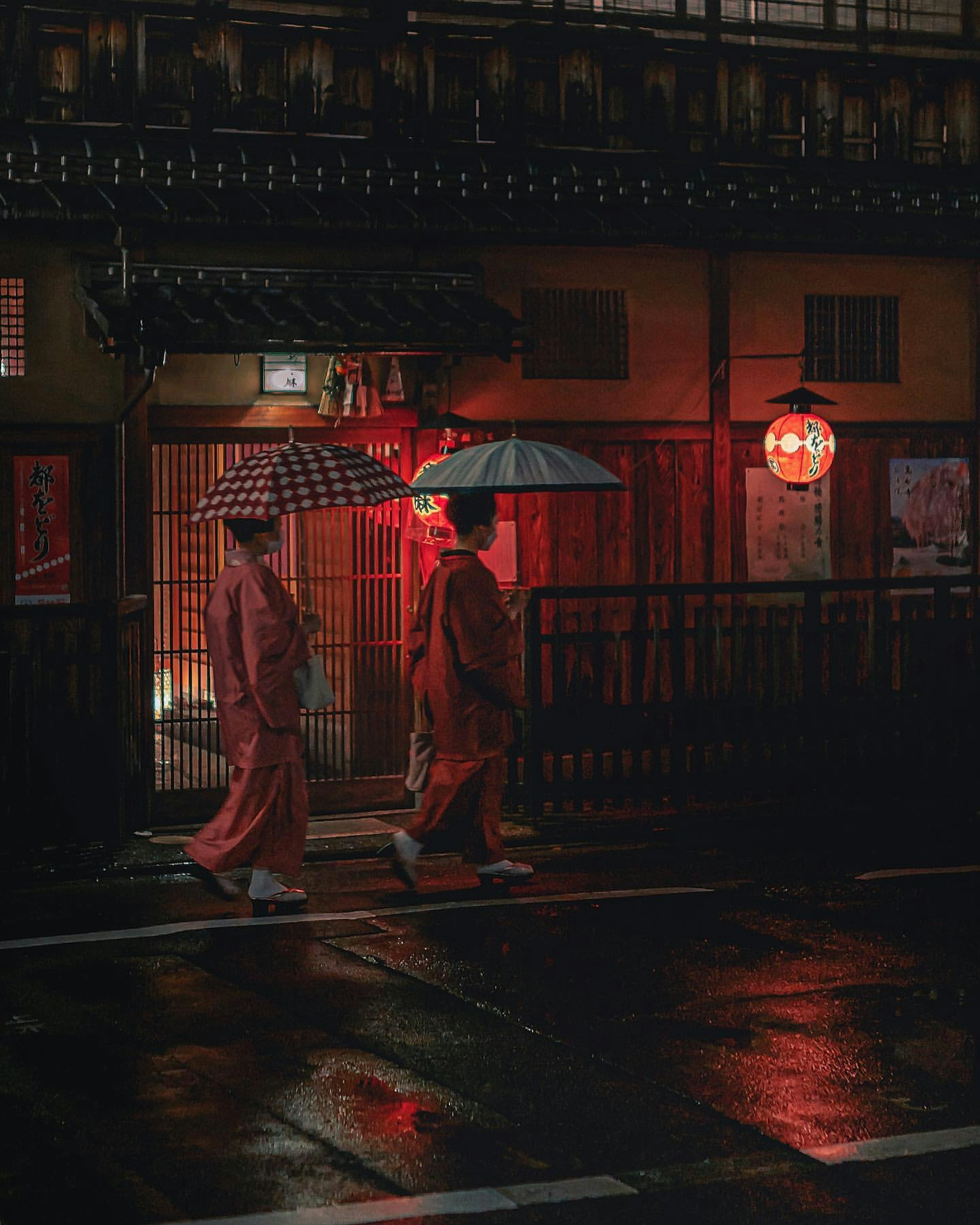 Zwei Frauen in roten Kimonos gehen mit Regenschirmen in einer regnerischen Straßenszene