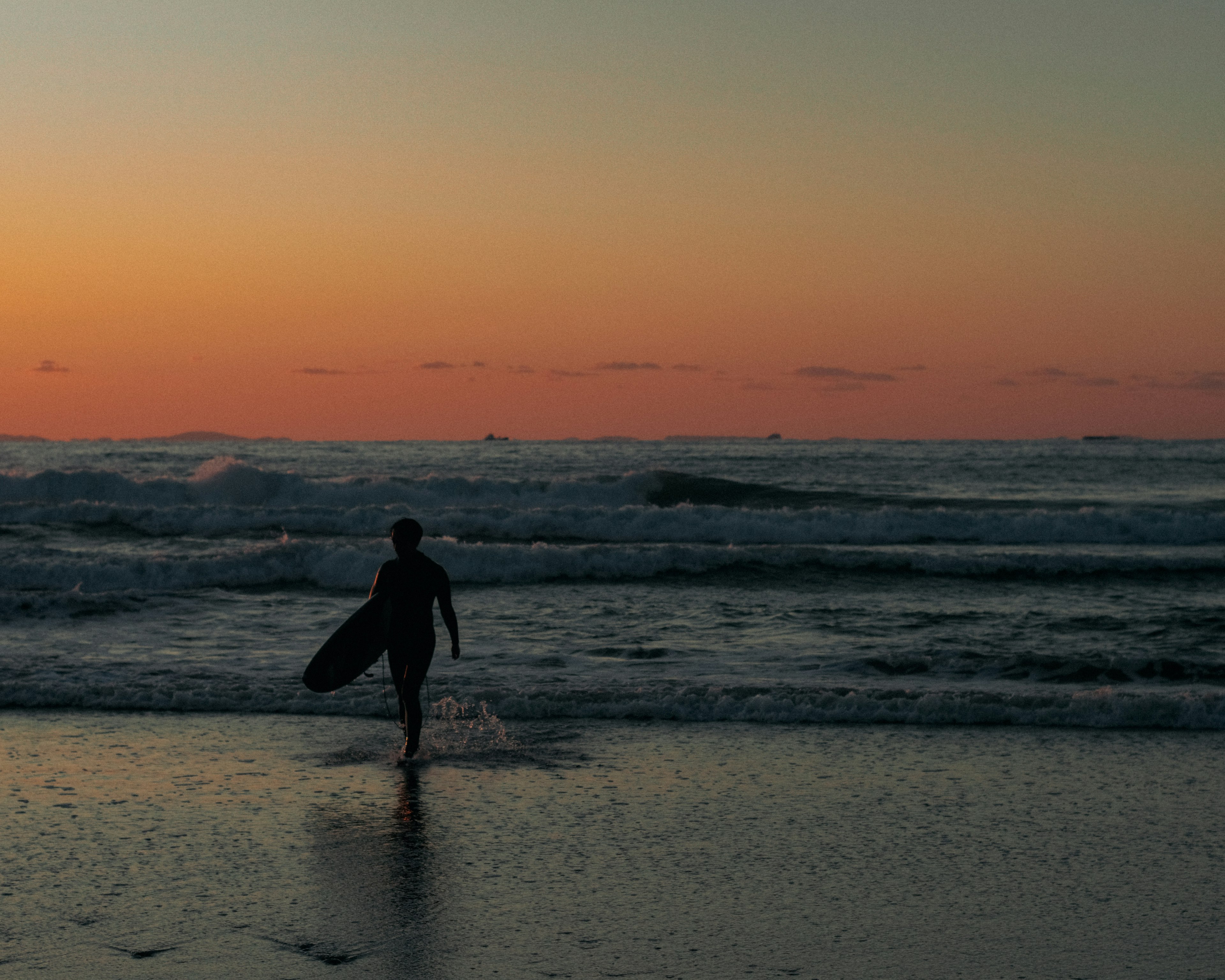 サーフボードを持ったシルエットのサーフィンをする人が波打つ海に向かう夕暮れのビーチ