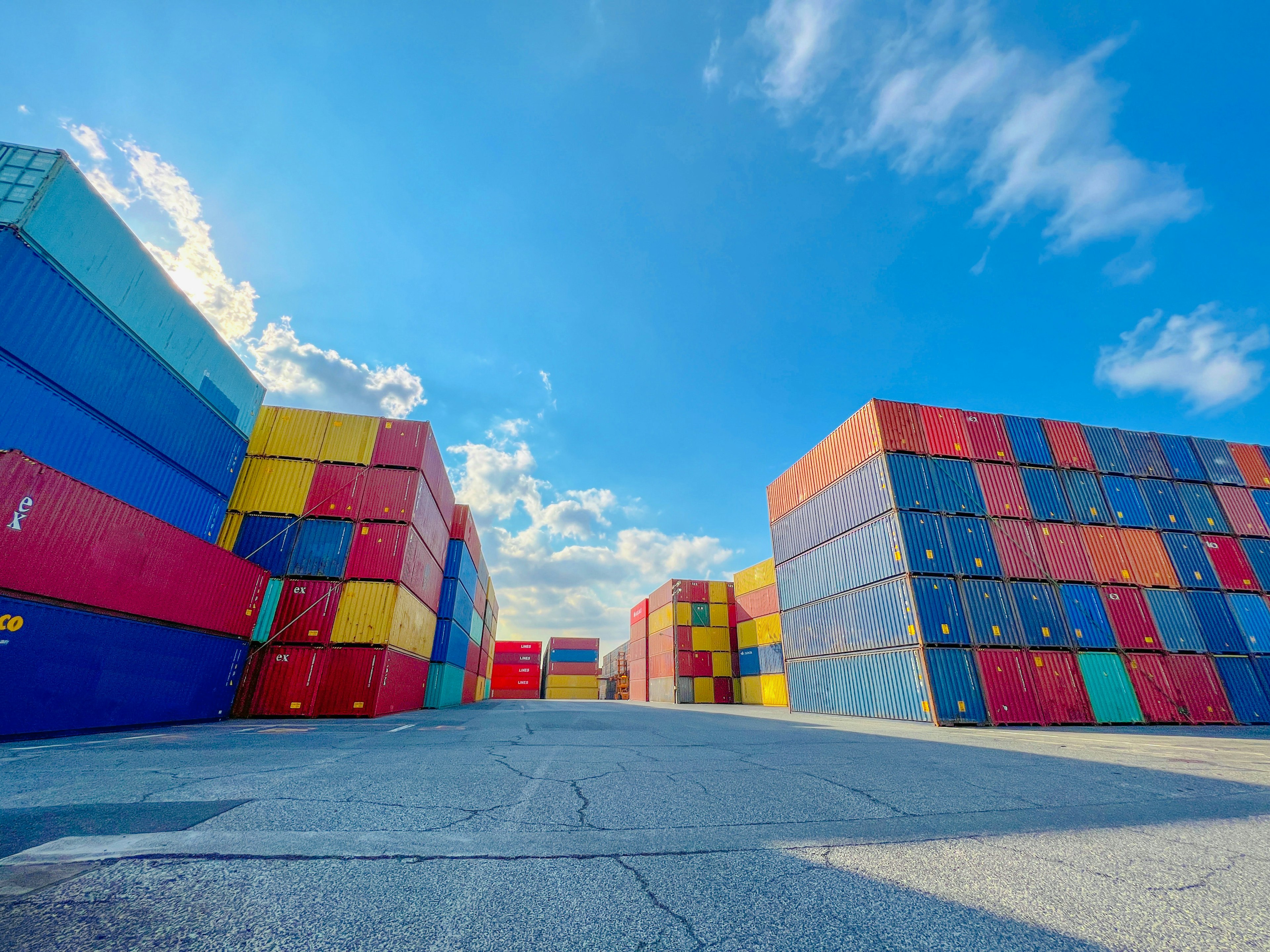 Colorful shipping containers stacked in a warehouse area under a blue sky with white clouds