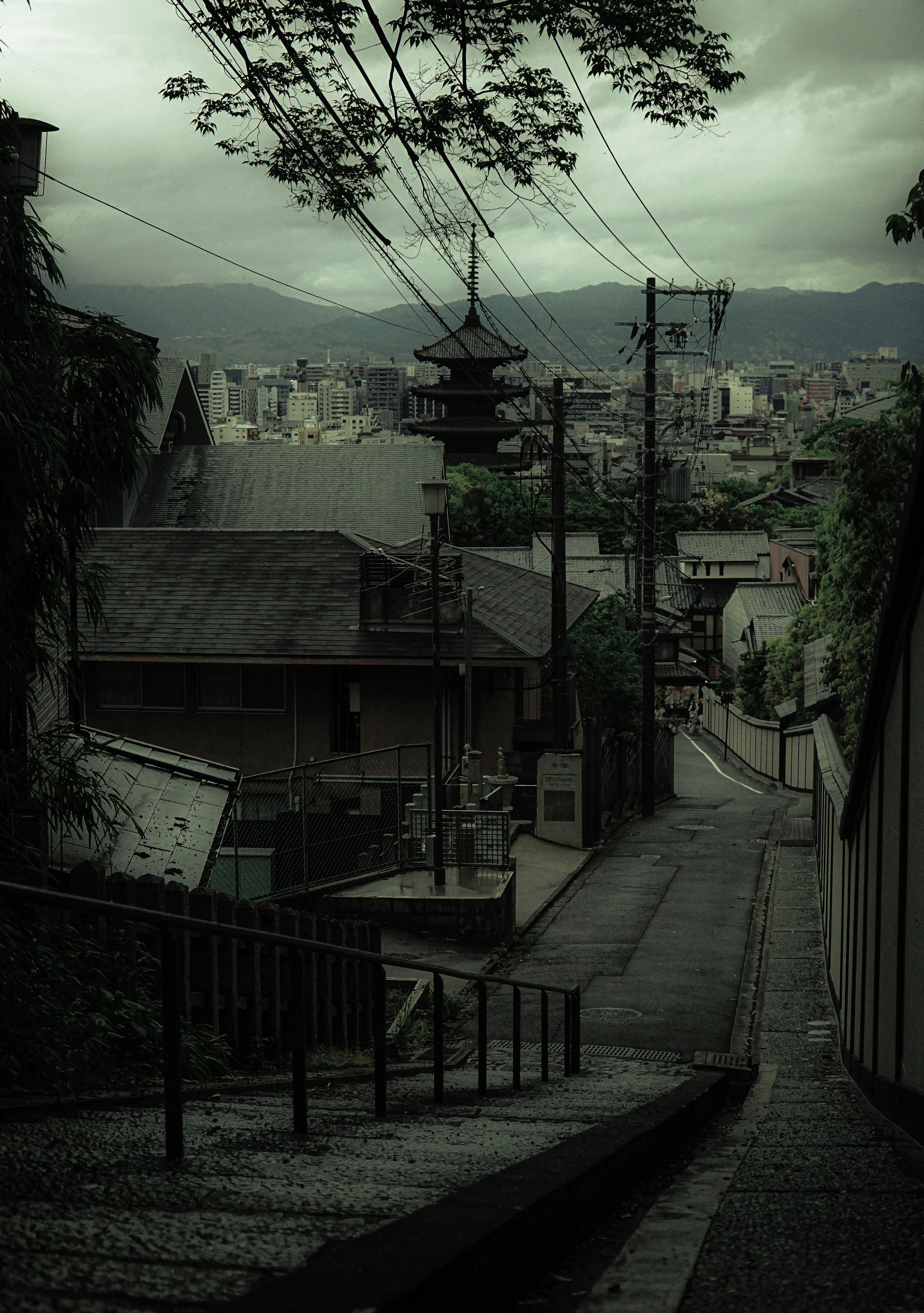 暗い雲に覆われた街並みと細い坂道が続く風景 古い建物と電柱が見える