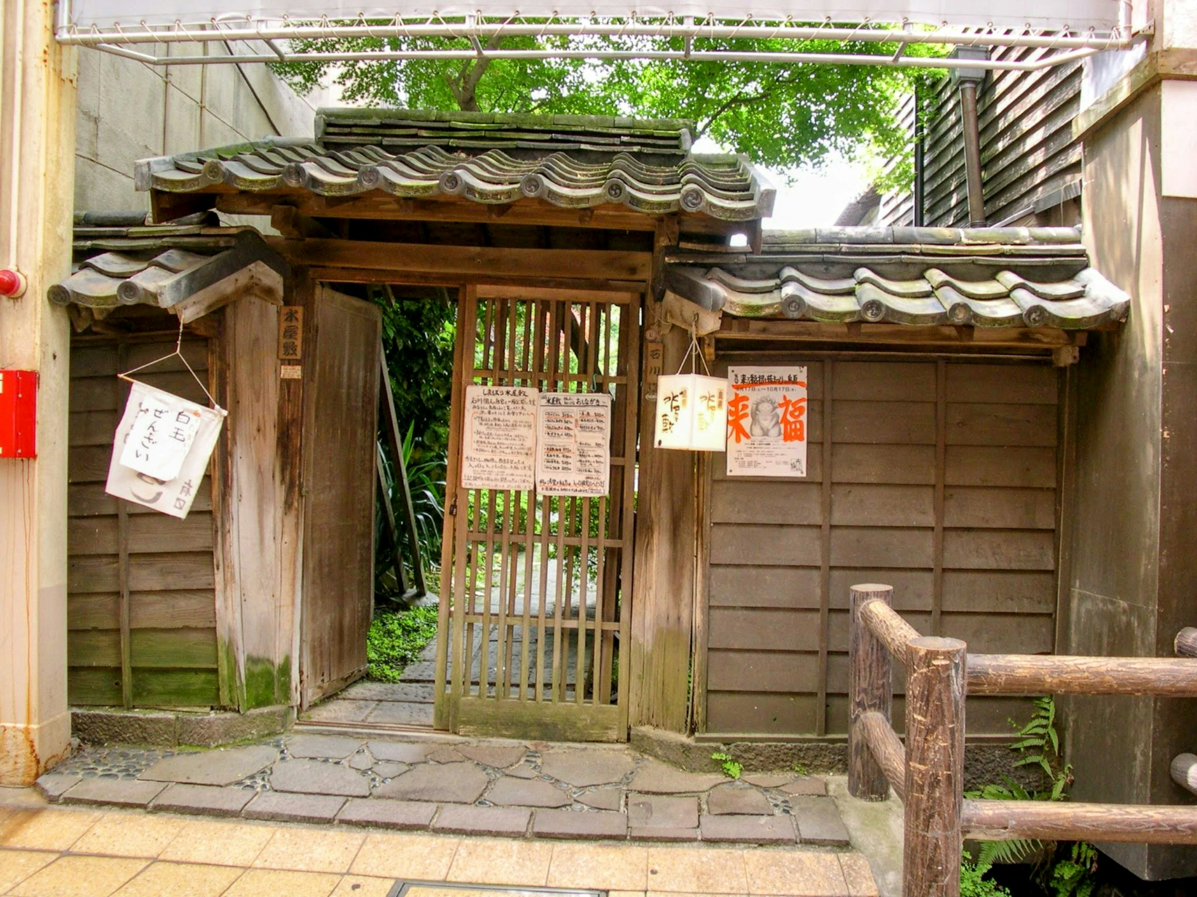 Exterior of an old house with a wooden gate and traditional Japanese roof surrounded by greenery